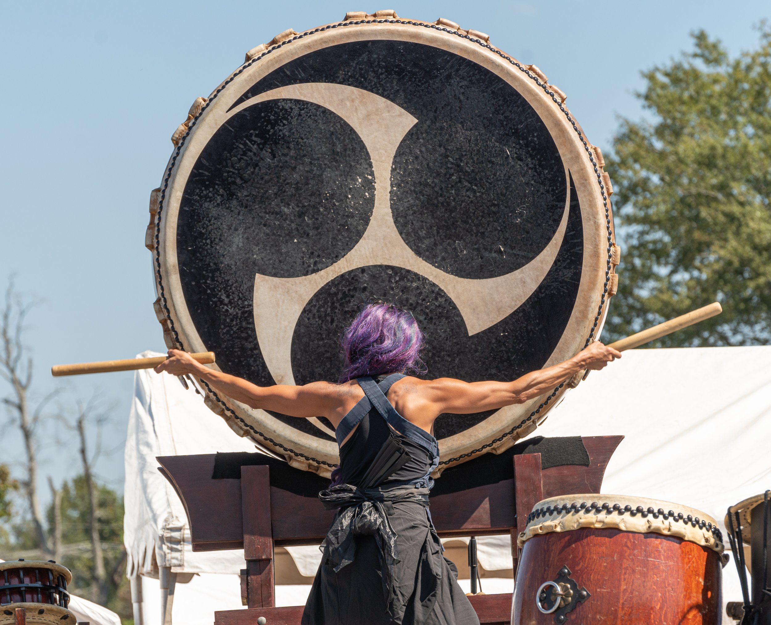 Person with purple hair wearing black clothing playing a large traditional drum outdoors, under a clear sky with trees in the background at the International Food & Art Festival