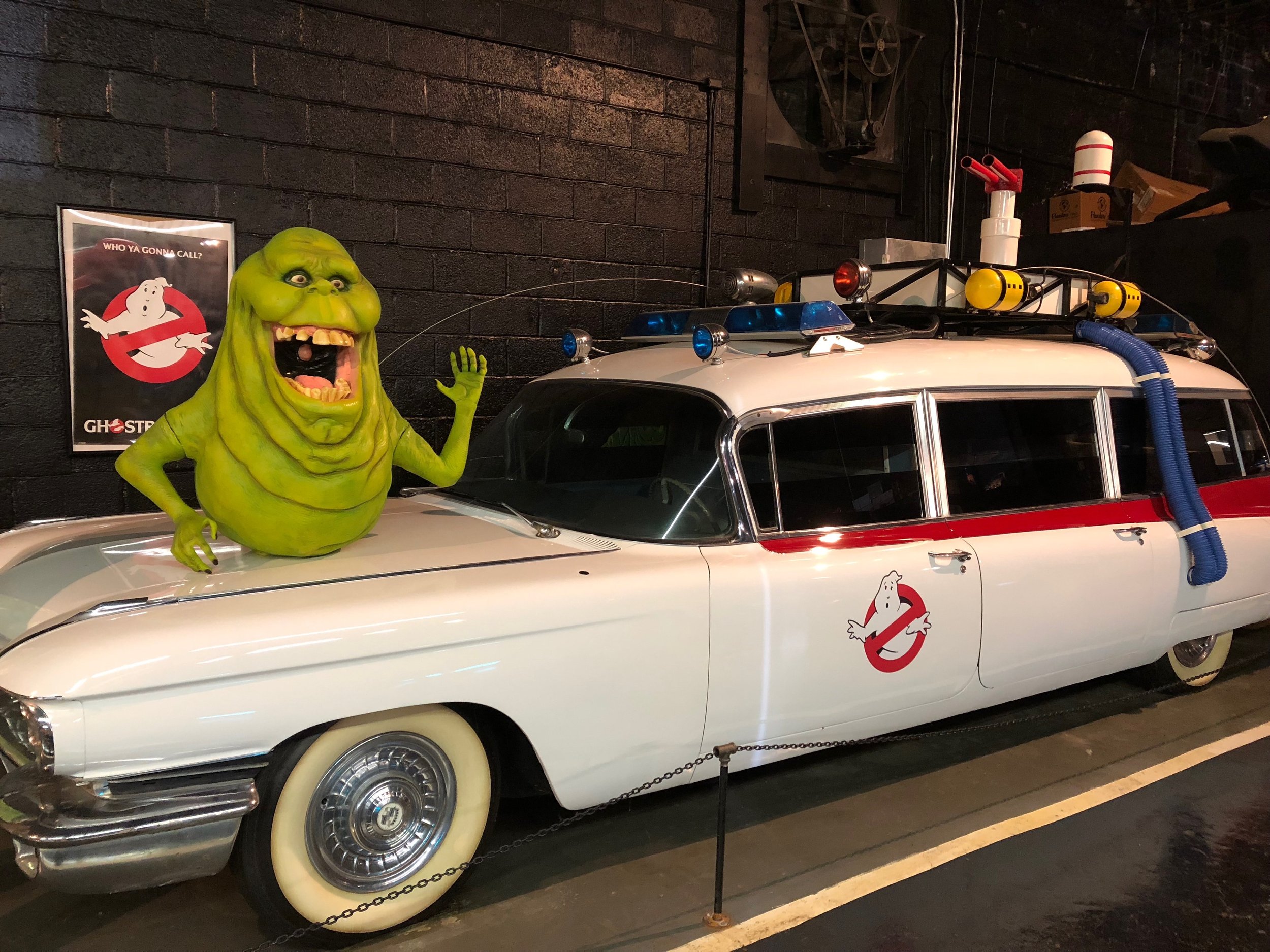 A vintage white Cadillac from Ghostbusters displayed at Rusty's TV & Movie Car Museum with a large green Ghostbusters character bust on its hood, and Ghostbusters-themed posters on the black brick wall behind it.