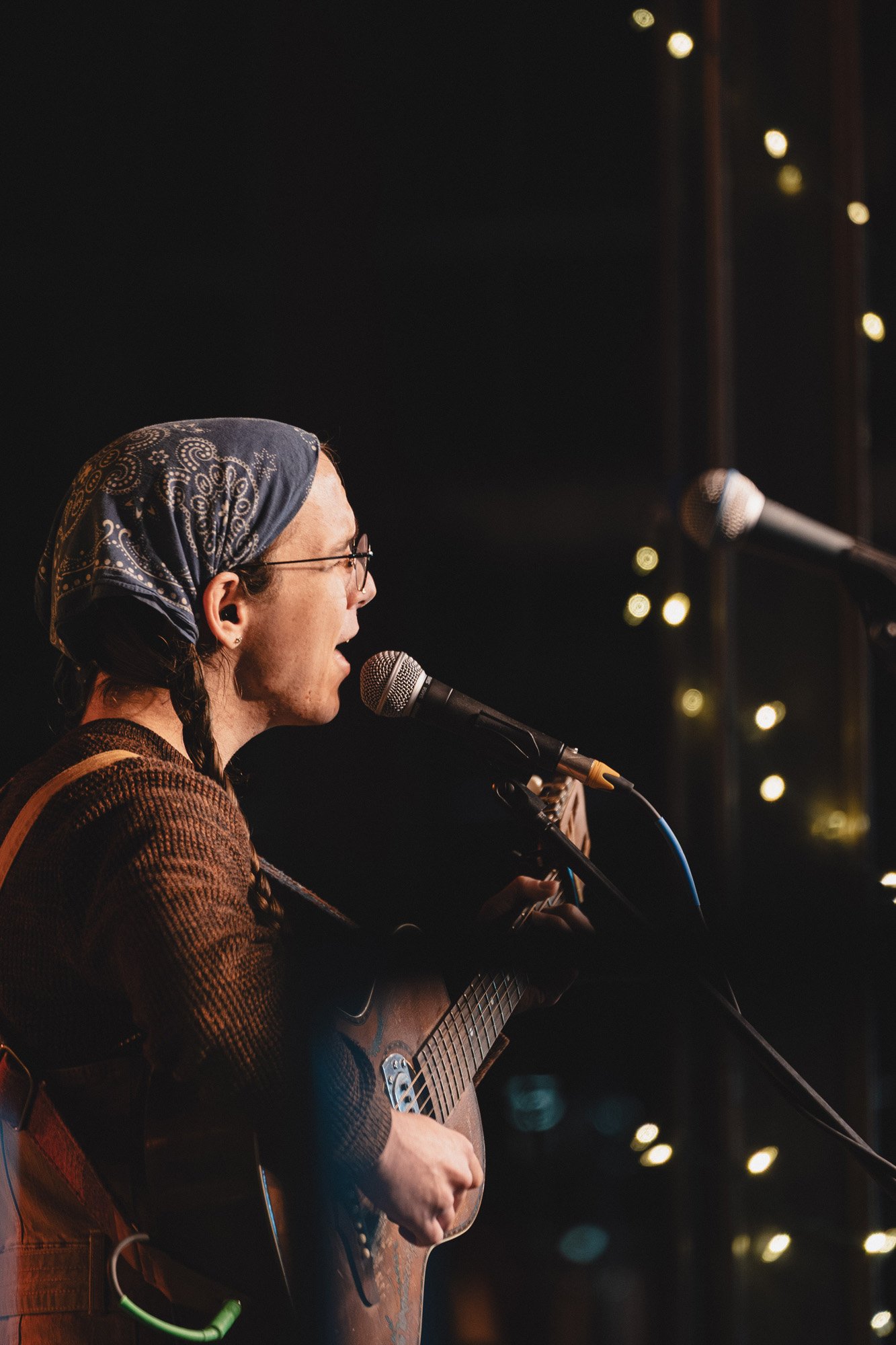 A woman wearing glasses and a bandana singing into a microphone, playing an acoustic guitar on stage with string lights in the background.