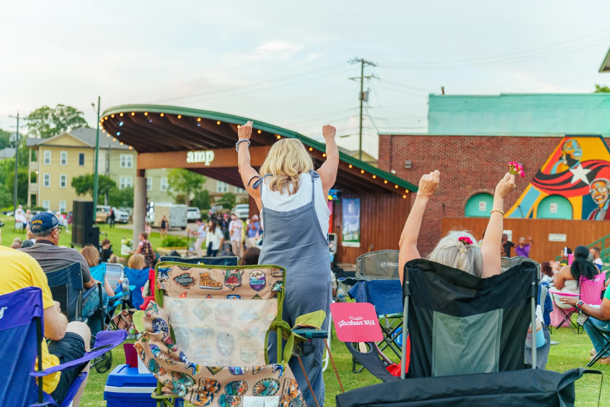 People seated in chairs outdoors at an AMP concert with colorful artwork in the background, and an older woman standing with arms raised, cheering.