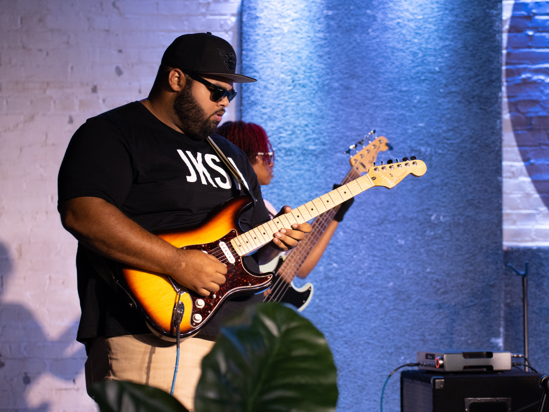 A man wearing sunglasses and a black cap playing an electric guitar during a music performance or rehearsal in an indoor setting with white brick walls and a blue textured wall.
