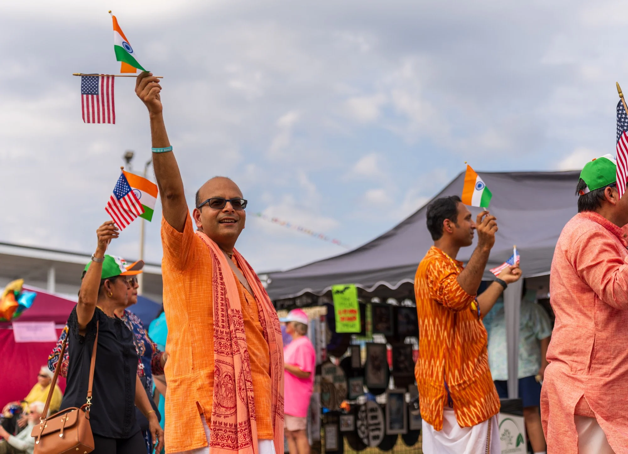 People celebrating India Independence Day, waving Indian flags and holding American flags at an outdoor event, with tents and a cloudy sky in the background.