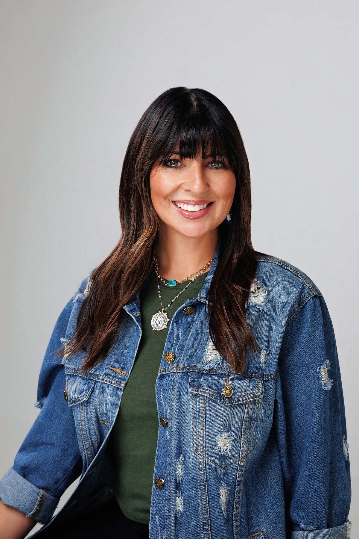 A woman with long dark hair and bangs, wearing a denim jacket with distressed details, a green t-shirt, and layered necklaces, smiling against a plain gray background.