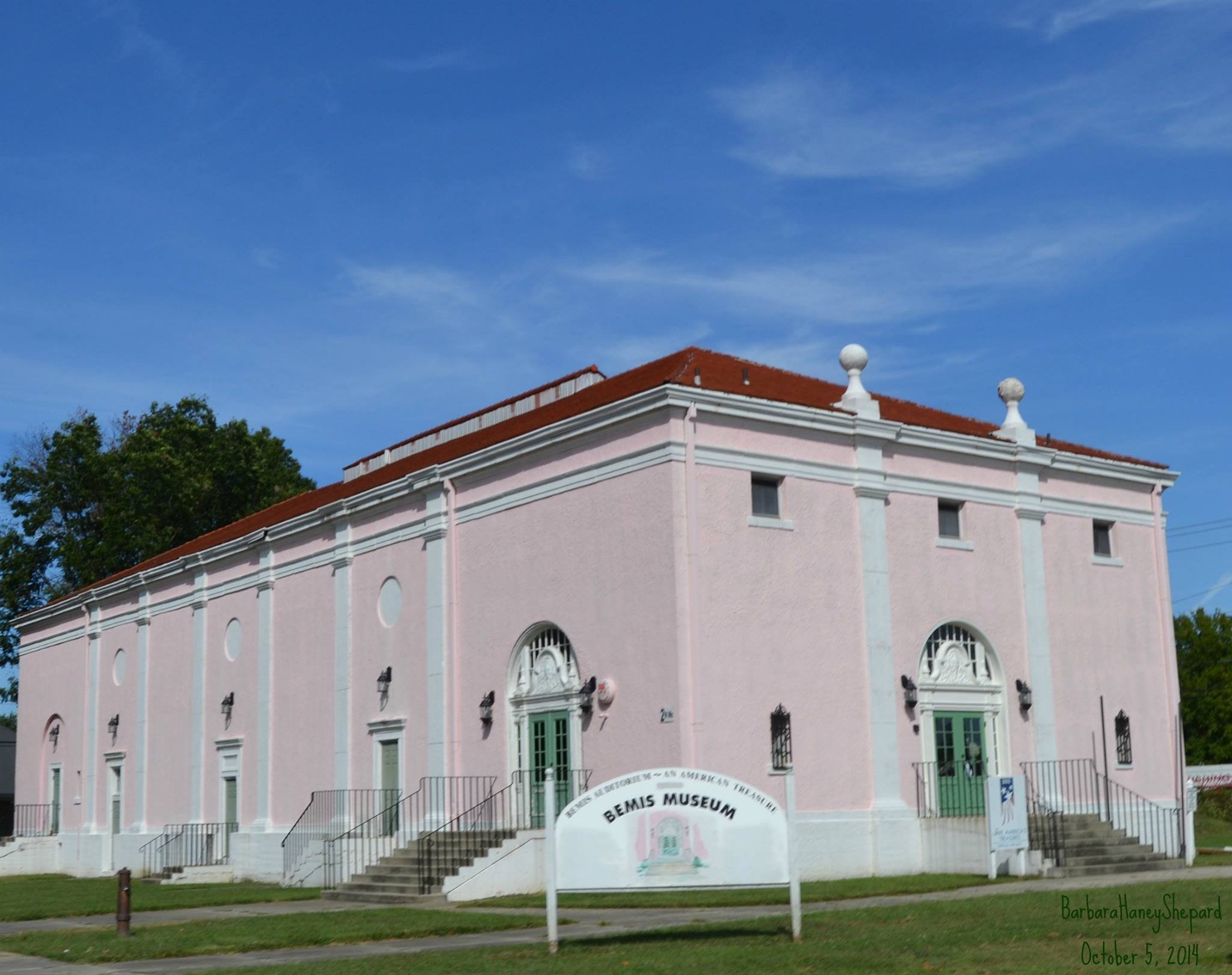Pink historic Bemis Mill with white accents, a red-tiled roof, and green doors, situated under a clear blue sky with some clouds. A sign reads 'Benis Museum' and there is an address and date on the bottom right corner.