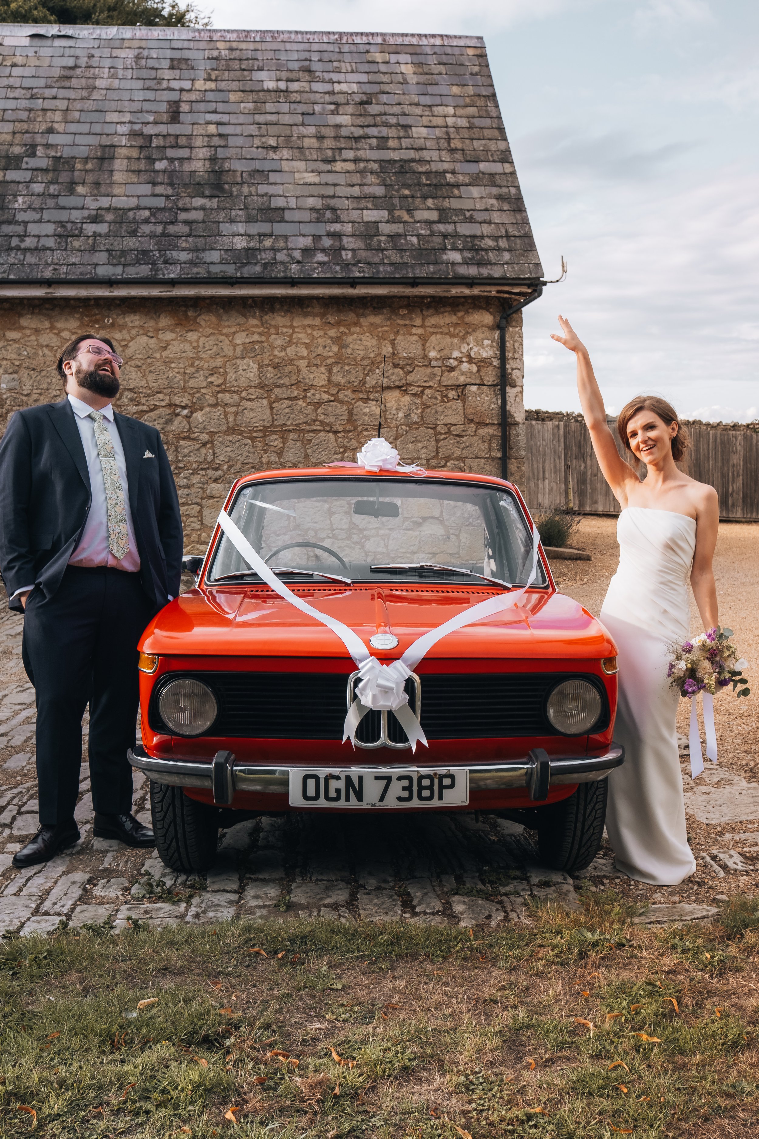 A bride and groom celebrating beside a vintage orange car decorated with white ribbons and a bow, outside a stone building, with the bride smiling and waving.