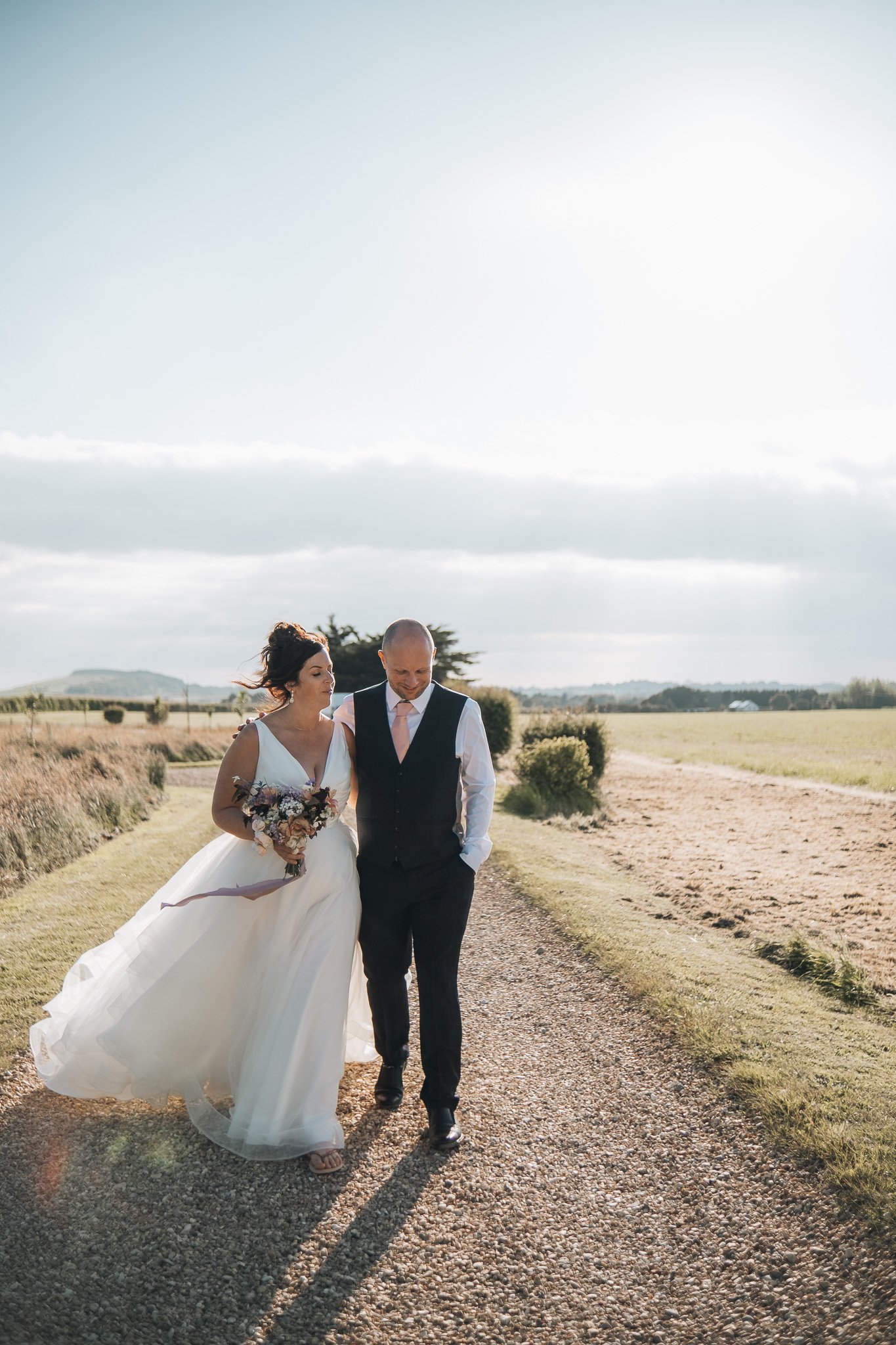 A bride in a white wedding gown and a groom in a vest and dress pants walk together on a dirt path outdoors in a rural setting, holding hands and smiling.