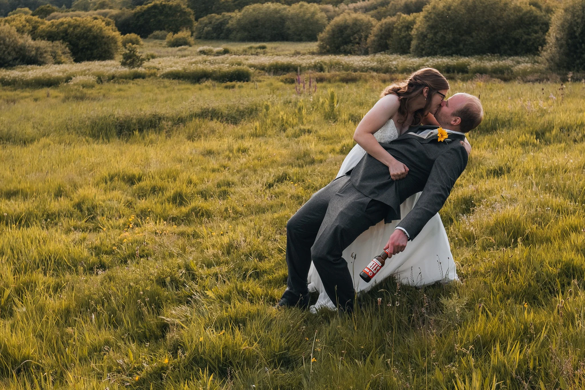 A bride and groom share a kiss in a grassy field with trees in the background, the groom is holding a beer and has a sunflower in his lapel.