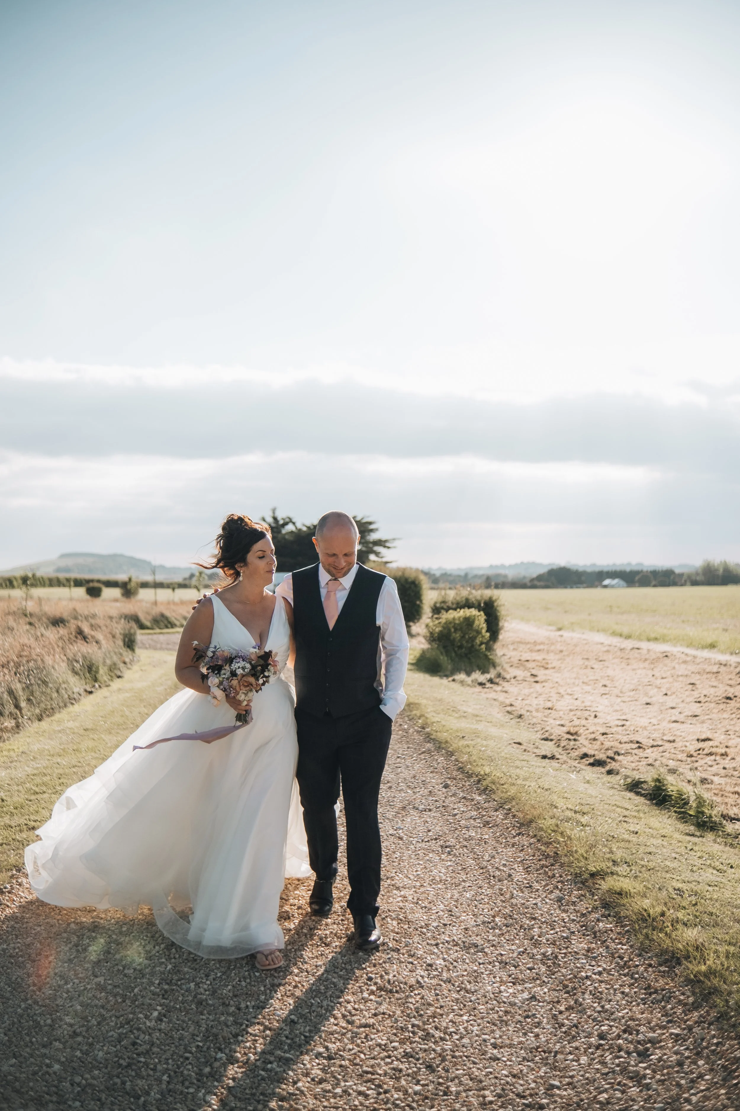 A bride and groom walking together on a gravel path outdoors, with open fields and a partly cloudy sky in the background, during their wedding celebration.