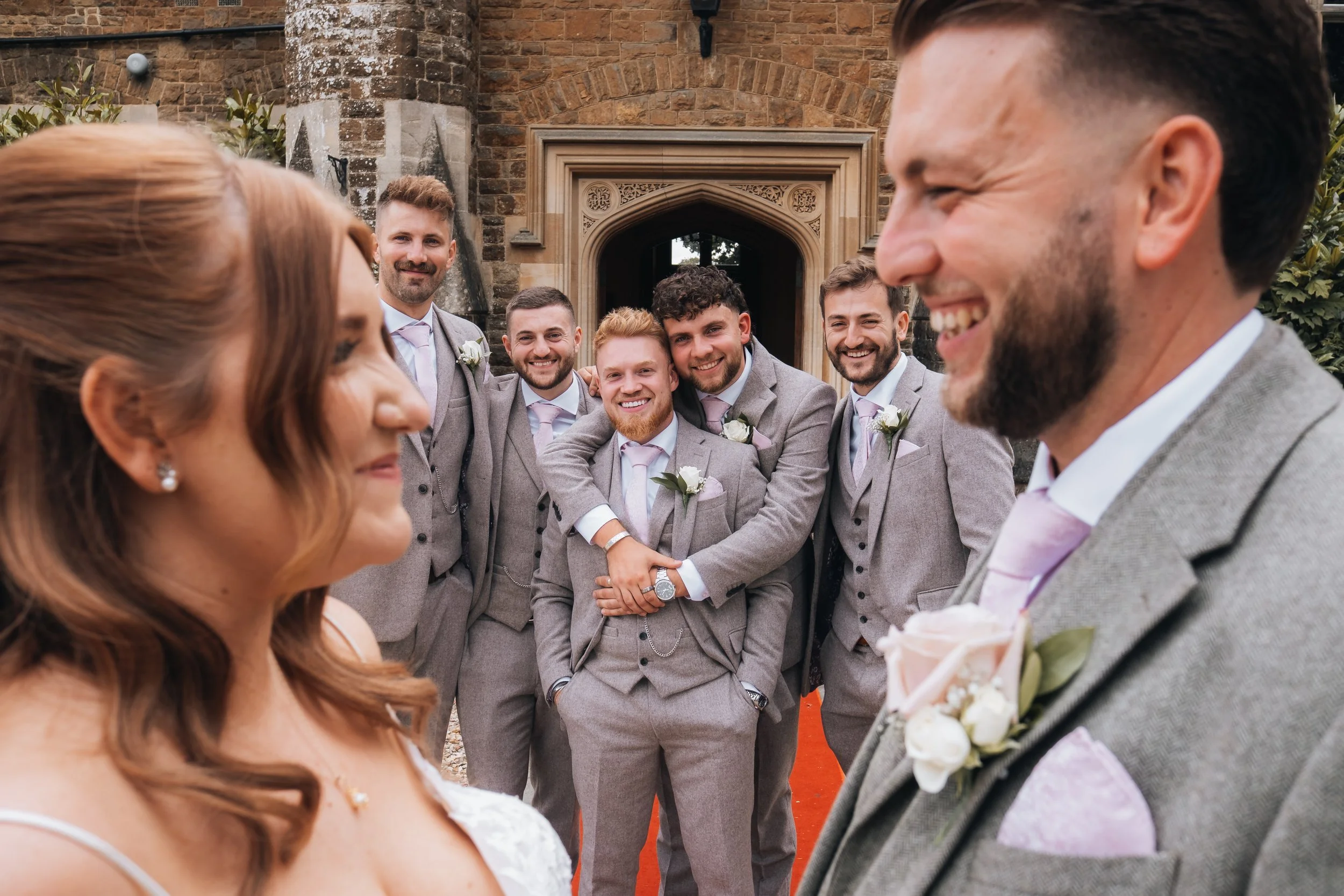 Bride and groom smiling and standing close together at the wedding ceremony, with the groom's groomsmen behind them, all dressed in matching gray suits with pink ties and floral boutonnieres. The setting appears to be outside a historic building with