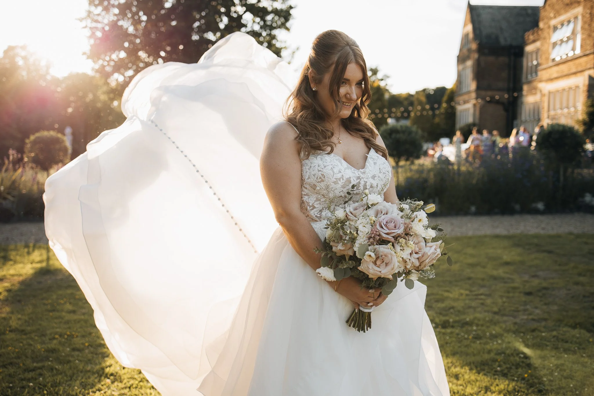 A bride in a white wedding dress holding a bouquet of pink and white roses, standing outdoors during sunset with greenery and a building in the background.