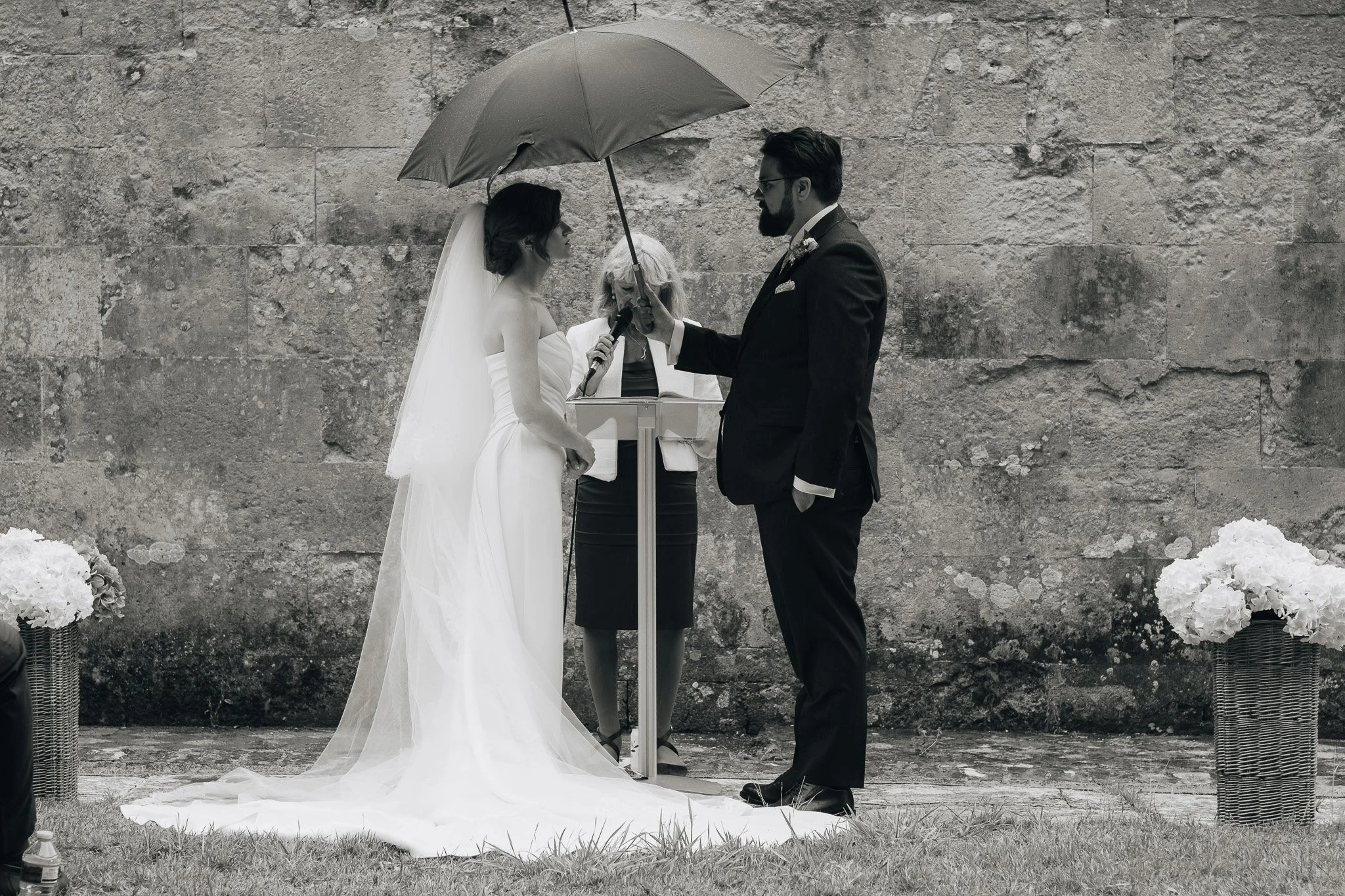 A couple getting married, standing outdoors under an umbrella, with an officiant in the background.