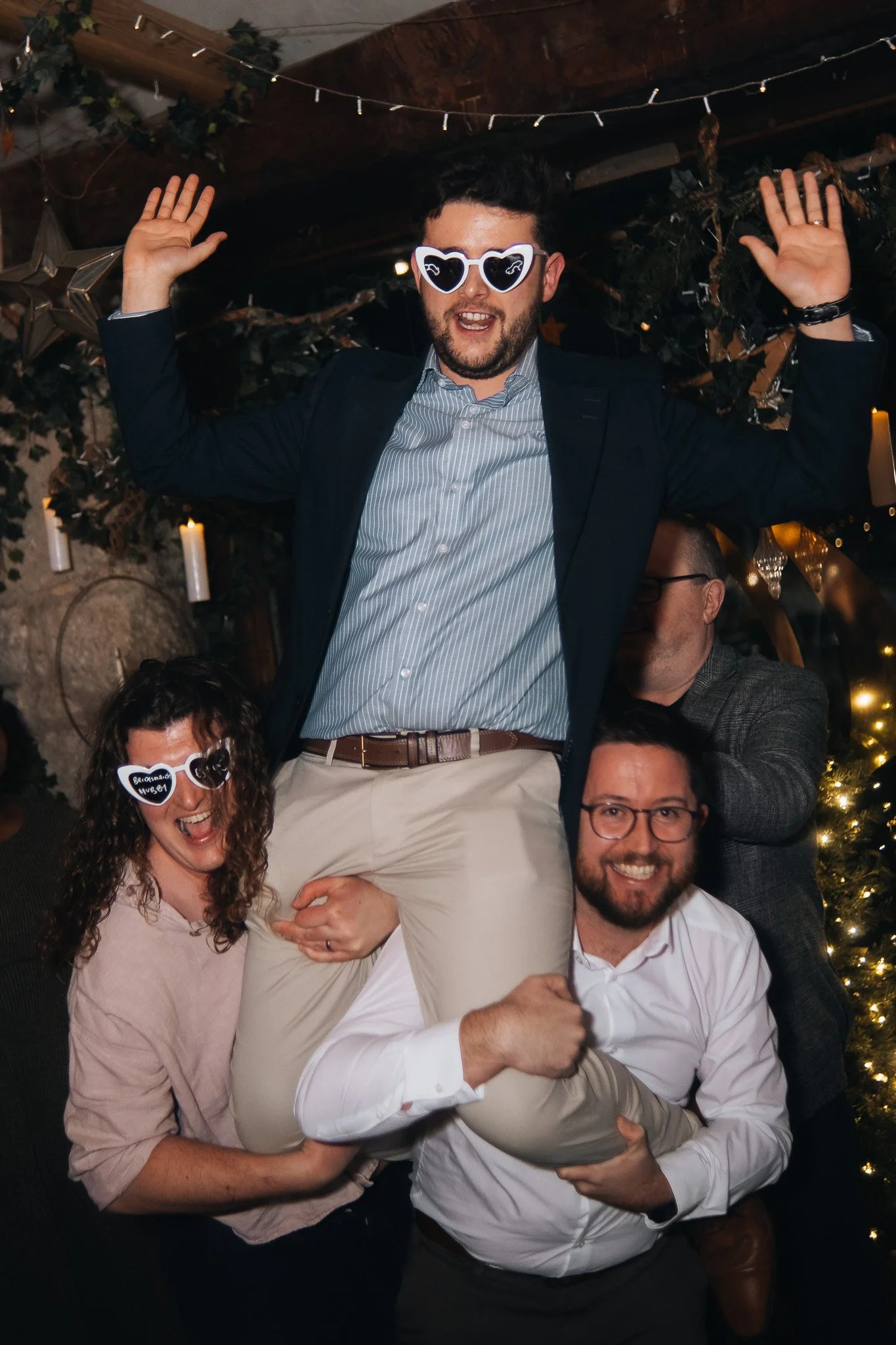 A group of five adults celebrating at a party, with one man wearing heart-shaped sunglasses and being lifted up by others. They are indoors with decorative string lights and candles.