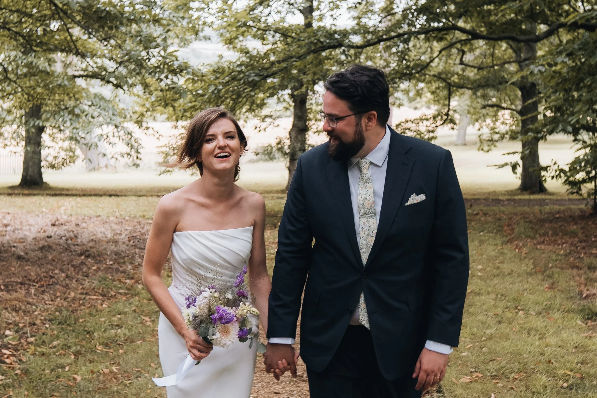 A bride and groom walking hand in hand outdoors, smiling, with trees and green grass in the background.