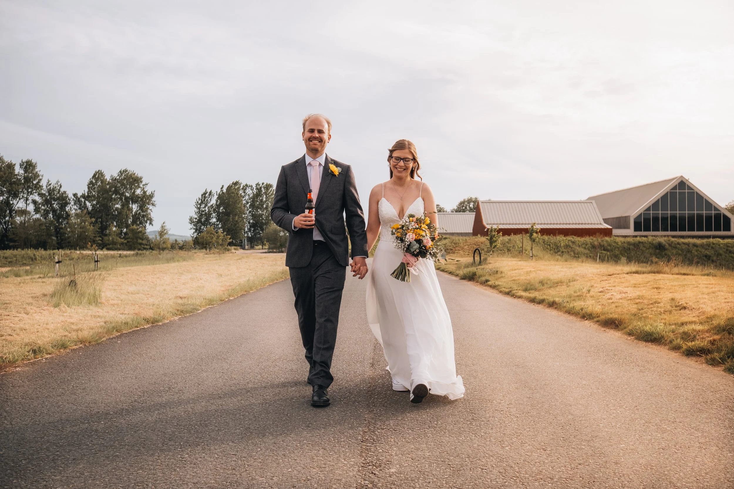 A newlywed couple walking hand in hand on a rural road during sunset, the bride holding a colorful bouquet and wearing a white wedding dress, the groom in a dark suit holding a beverage.