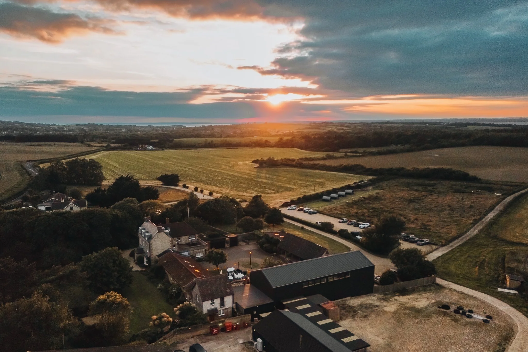 Aerial view of a rural landscape at sunset, featuring houses, a parking lot with cars, green fields, and a cloudy sky.