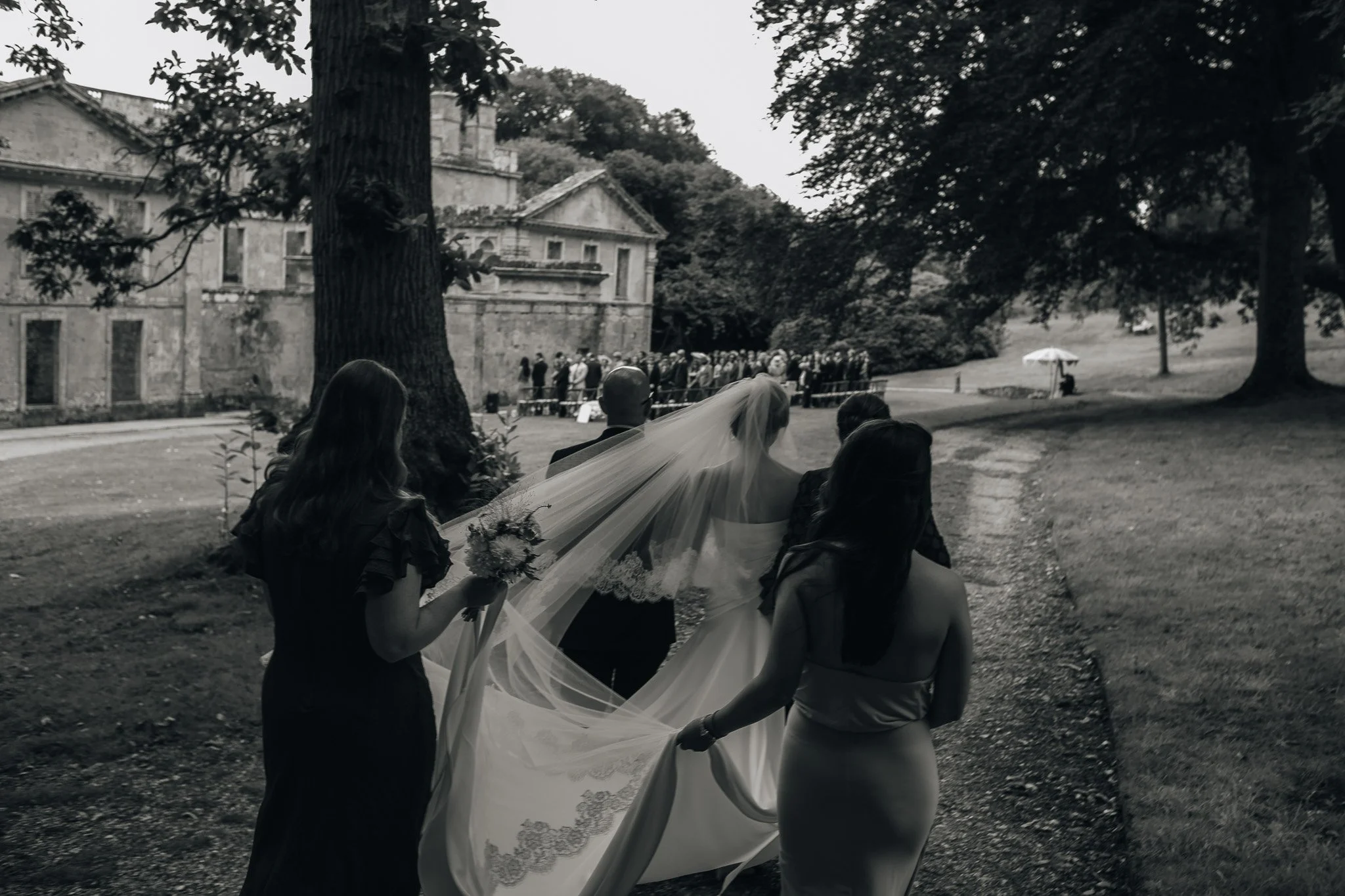 A wedding procession outdoors featuring a bride with a long veil, walking with three women, one holding her train, in front of a large historic building.