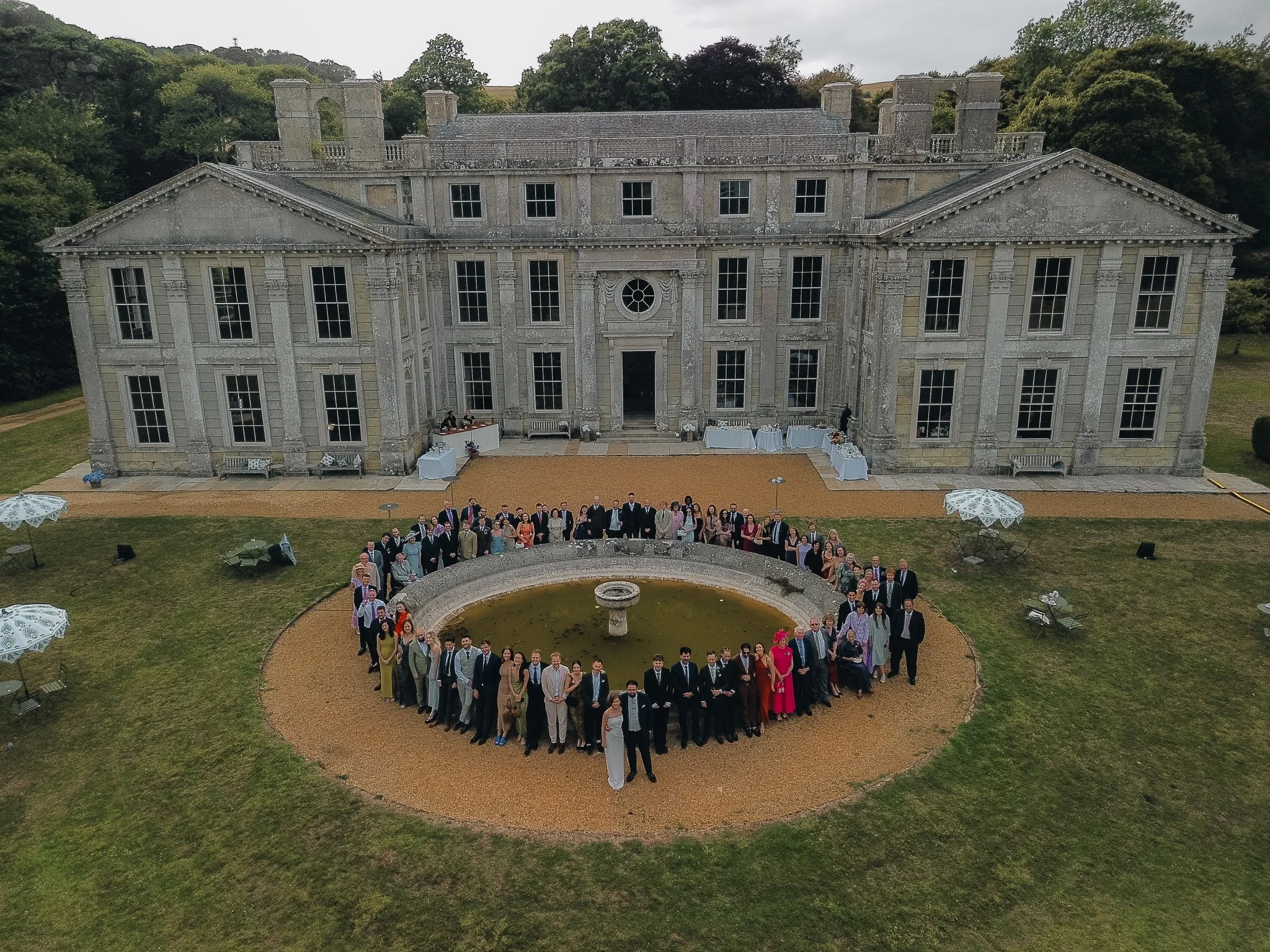 An aerial view of a large group of people posing in front of a historic mansion with stone exterior, tall windows, and classical architecture, surrounded by a well-maintained lawn and a circular fountain.