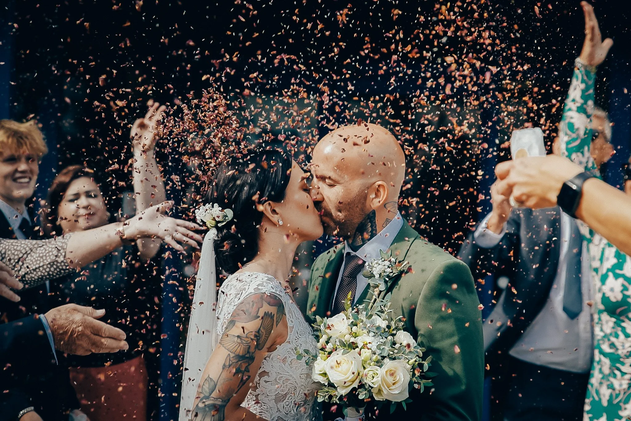 A couple getting married, kissing, with confetti falling around them during a celebration with friends.