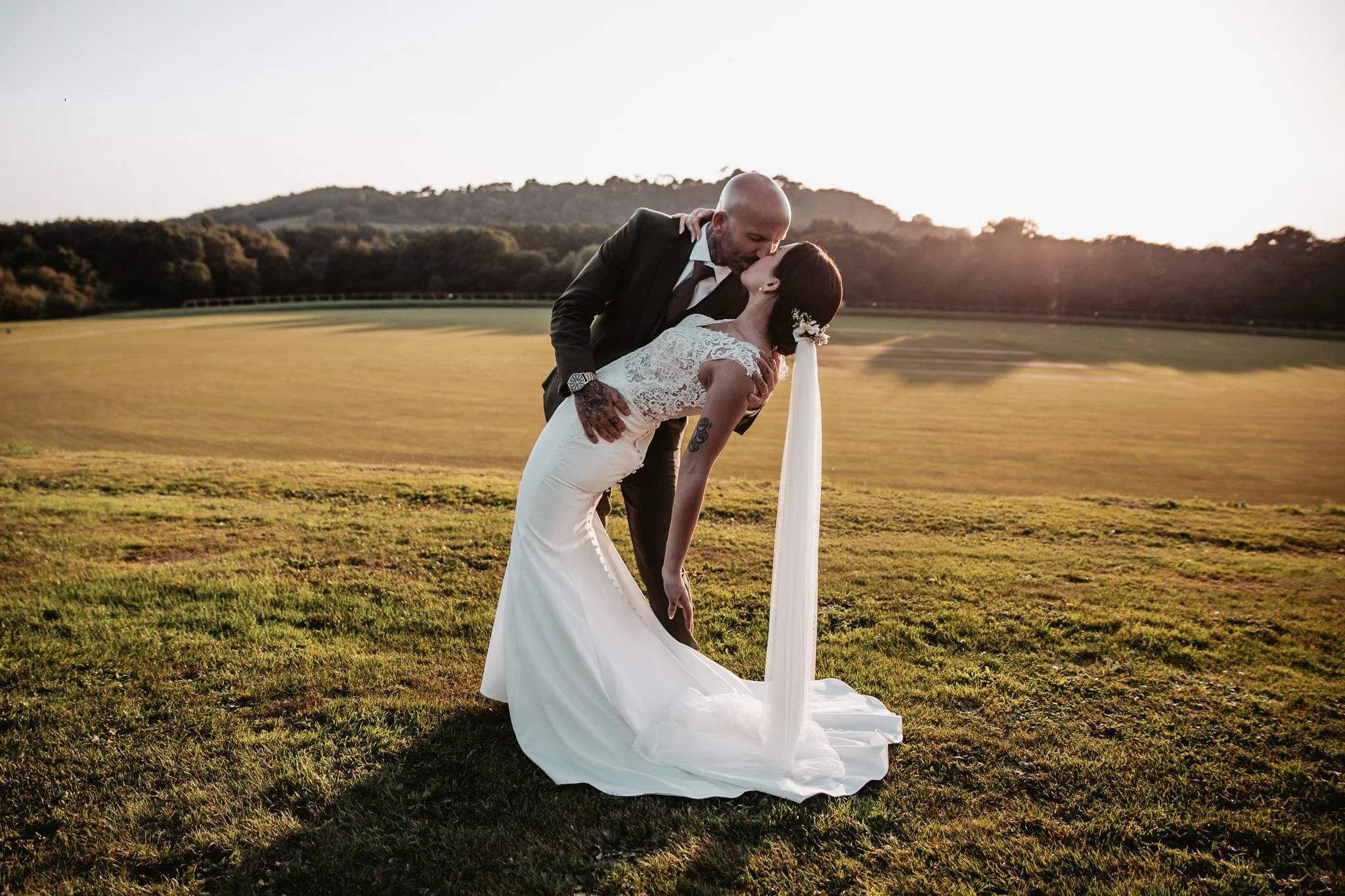 A bride and groom kiss outdoors on a grassy field during sunset, with the bride leaning back in the groom's arms.