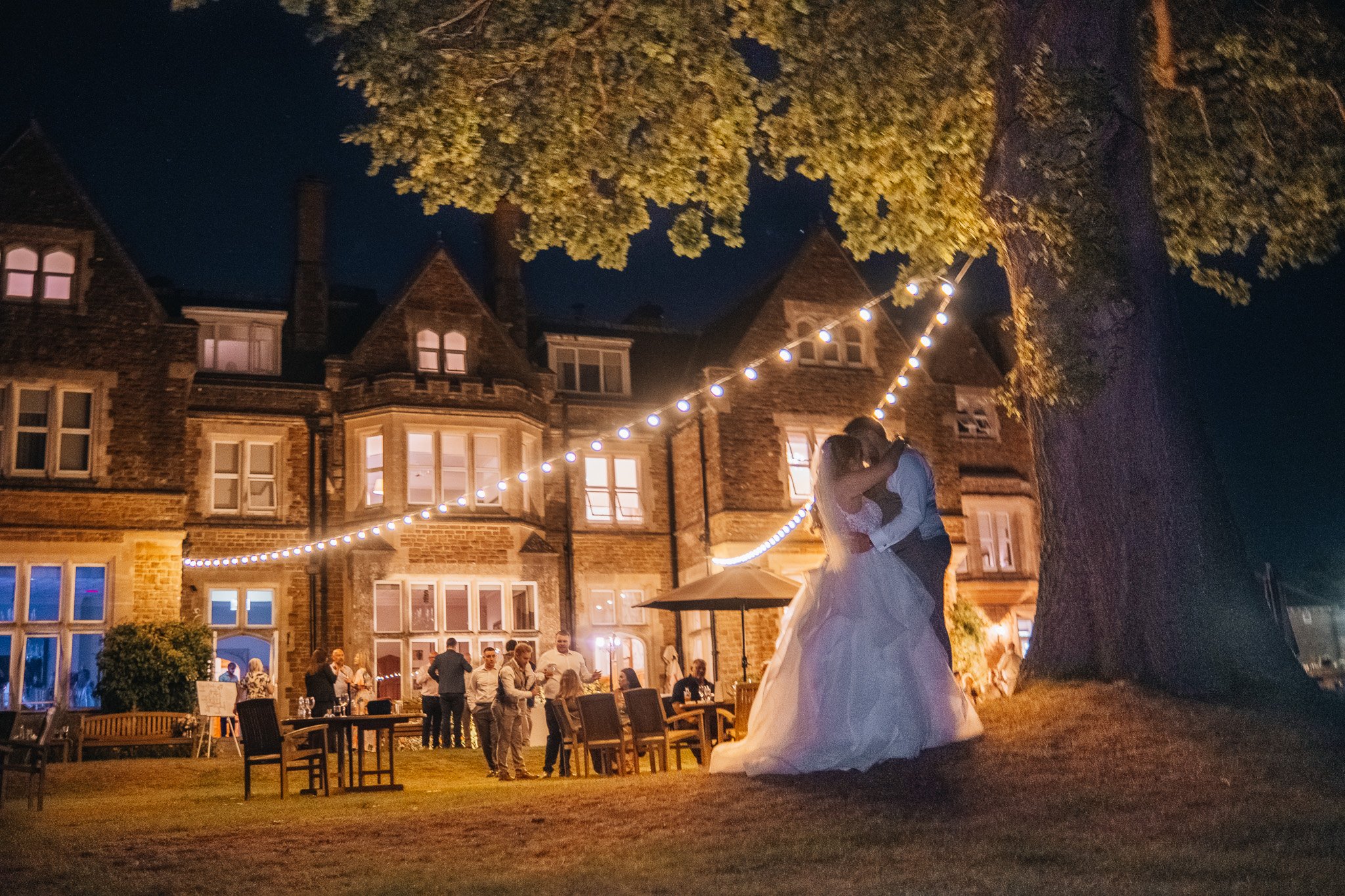 A bride and groom dancing under string lights outside a large brick house at night, with guests mingling in the background.