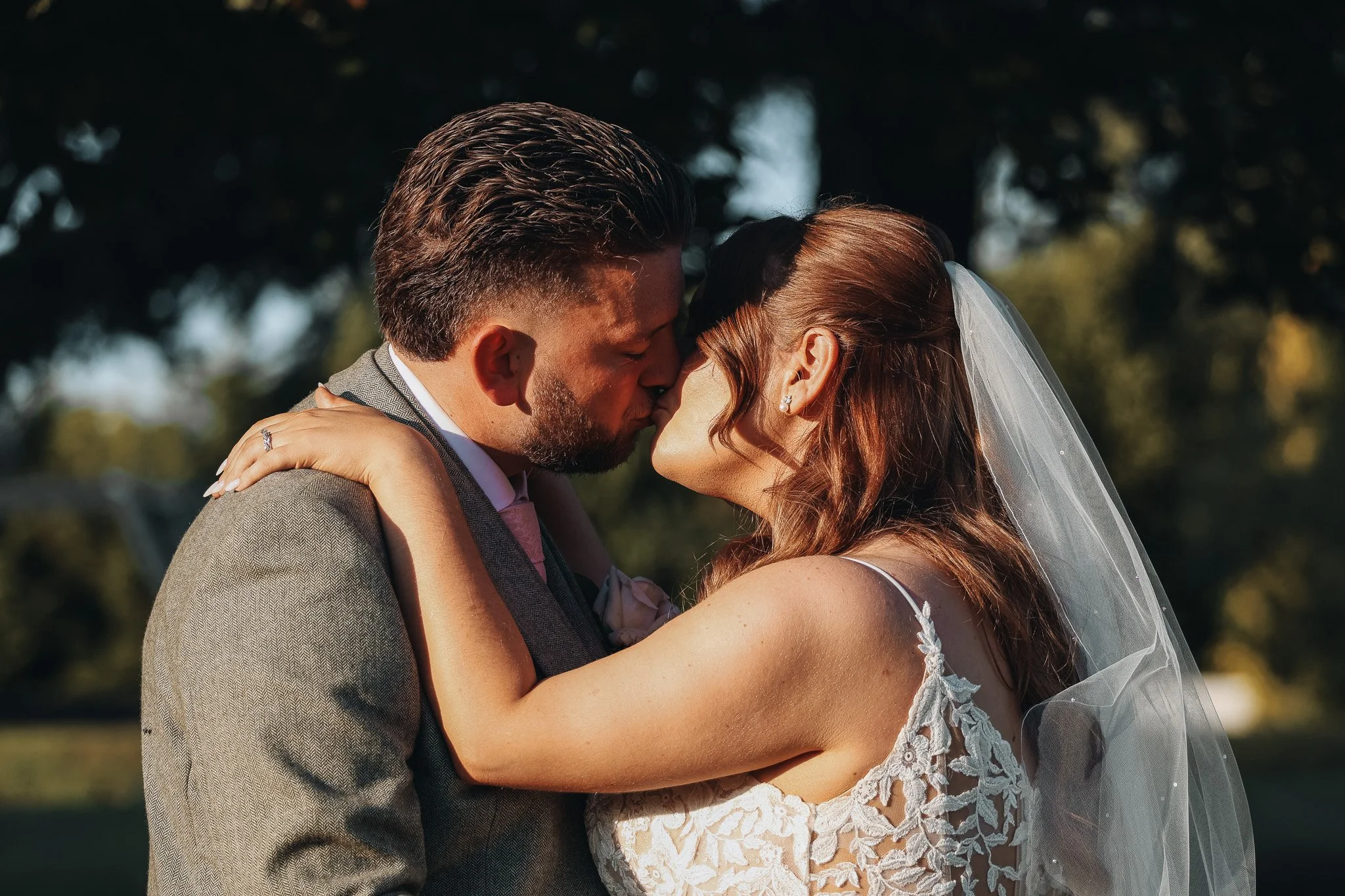 A wedding couple shares a kiss outdoors, with trees in the background. The groom is wearing a gray suit, and the bride is in a white lace wedding dress with a veil.
