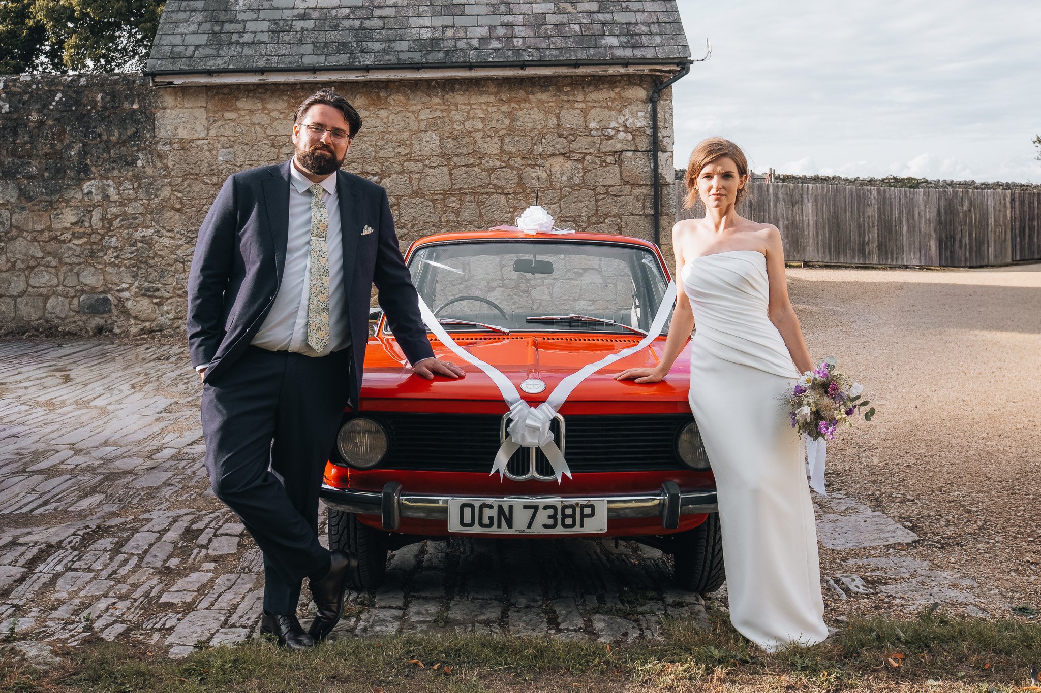 A bride and groom standing beside a red vintage car decorated with white ribbons and a bow, outside near a stone wall and fence.