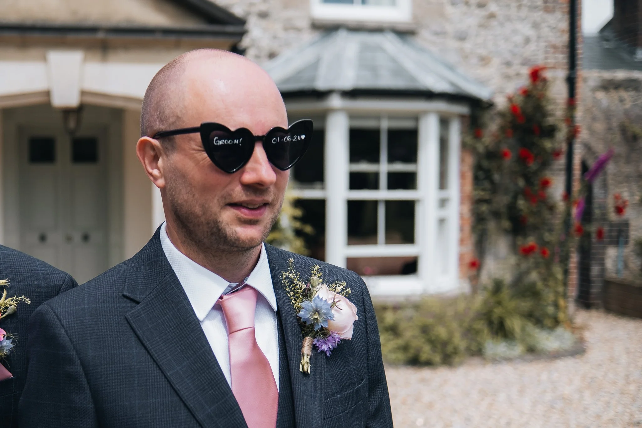A man wearing sunglasses with writing on the lenses, a dark checkered suit, a pink tie, and a boutonniere, standing outdoors in front of a building with a white window frame and some greenery.