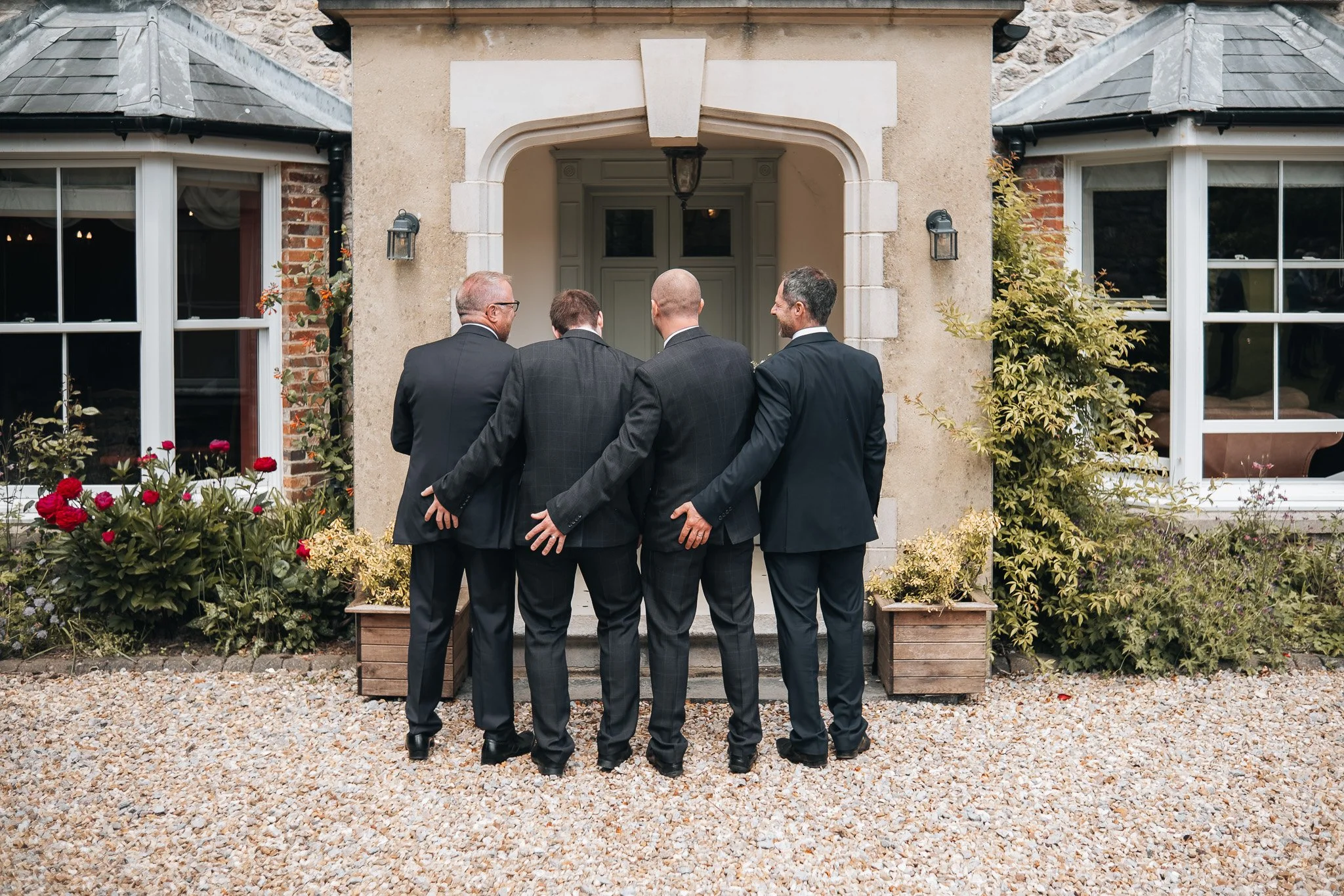 Four men dressed in suits standing close together in front of a house, seen from behind.