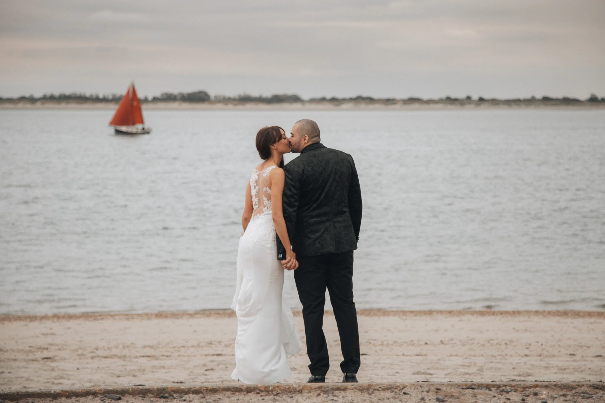 A bride and groom in wedding attire kissing on a beach near a body of water, with a sailing boat with red sails in the background, during overcast weather.