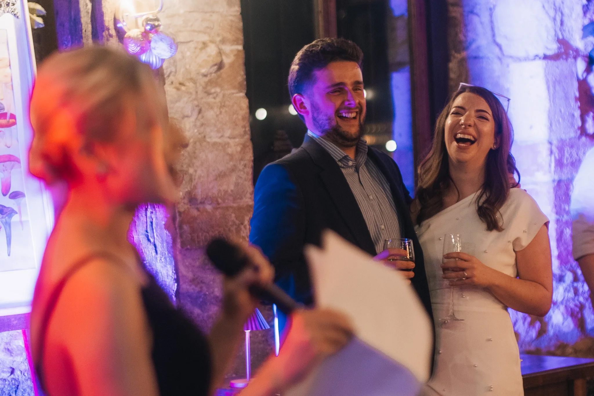 A woman giving a speech with a microphone at a celebration or wedding reception, with guests in the background, including a man and woman who are smiling and holding drinks.