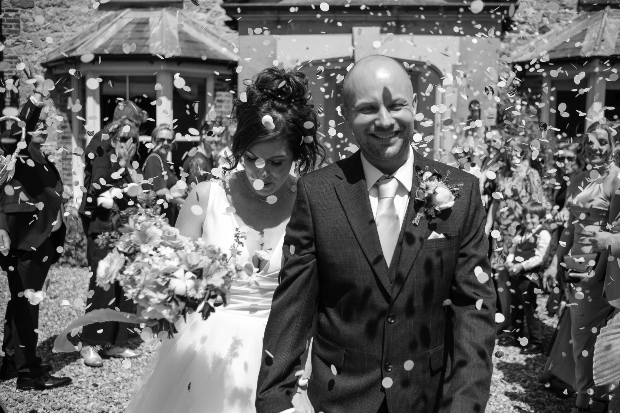 Black-and-white photo of a wedding celebration with a smiling groom in a suit and a bride carrying a bouquet, surrounded by guests throwing confetti.