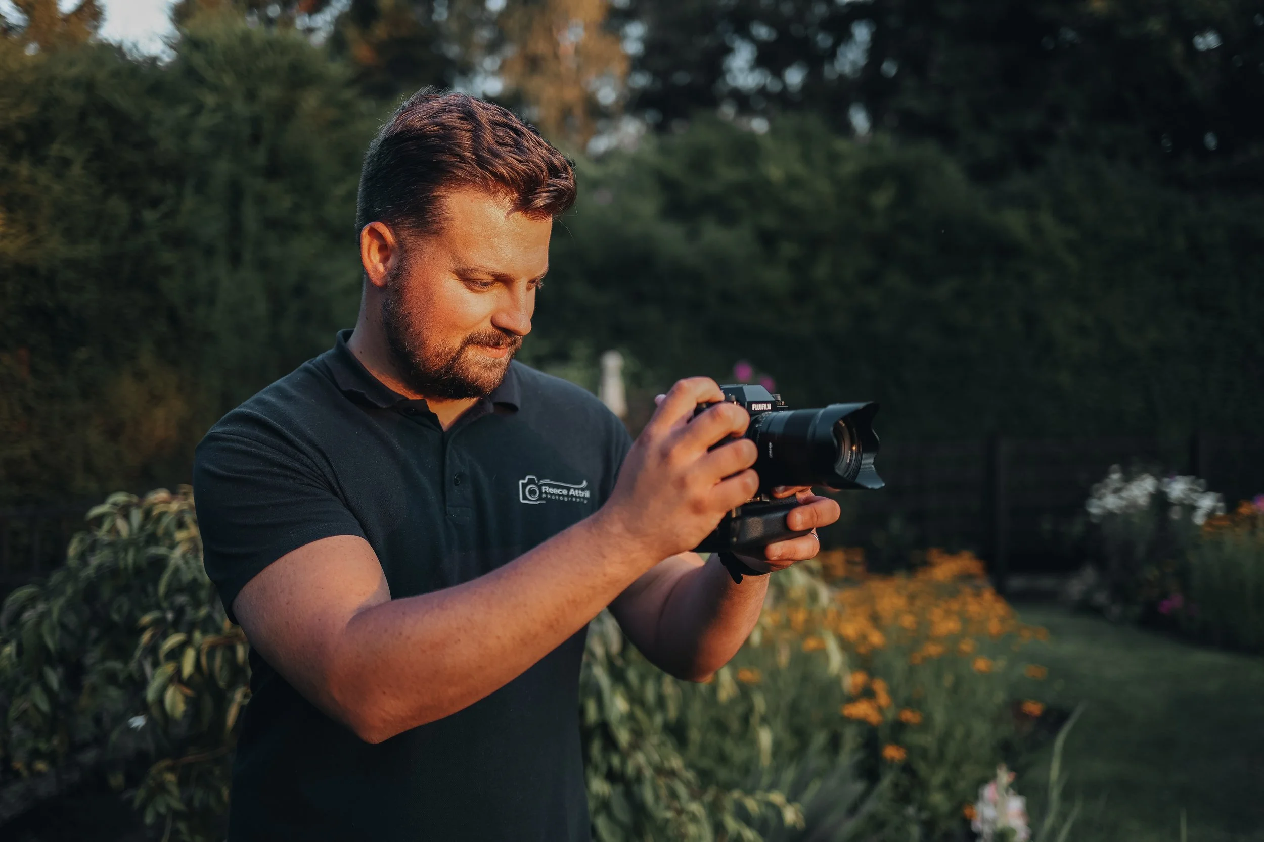 A man with a beard and wearing a black polo shirt with a camera logo, holding a professional camera outdoors during sunset in a garden.