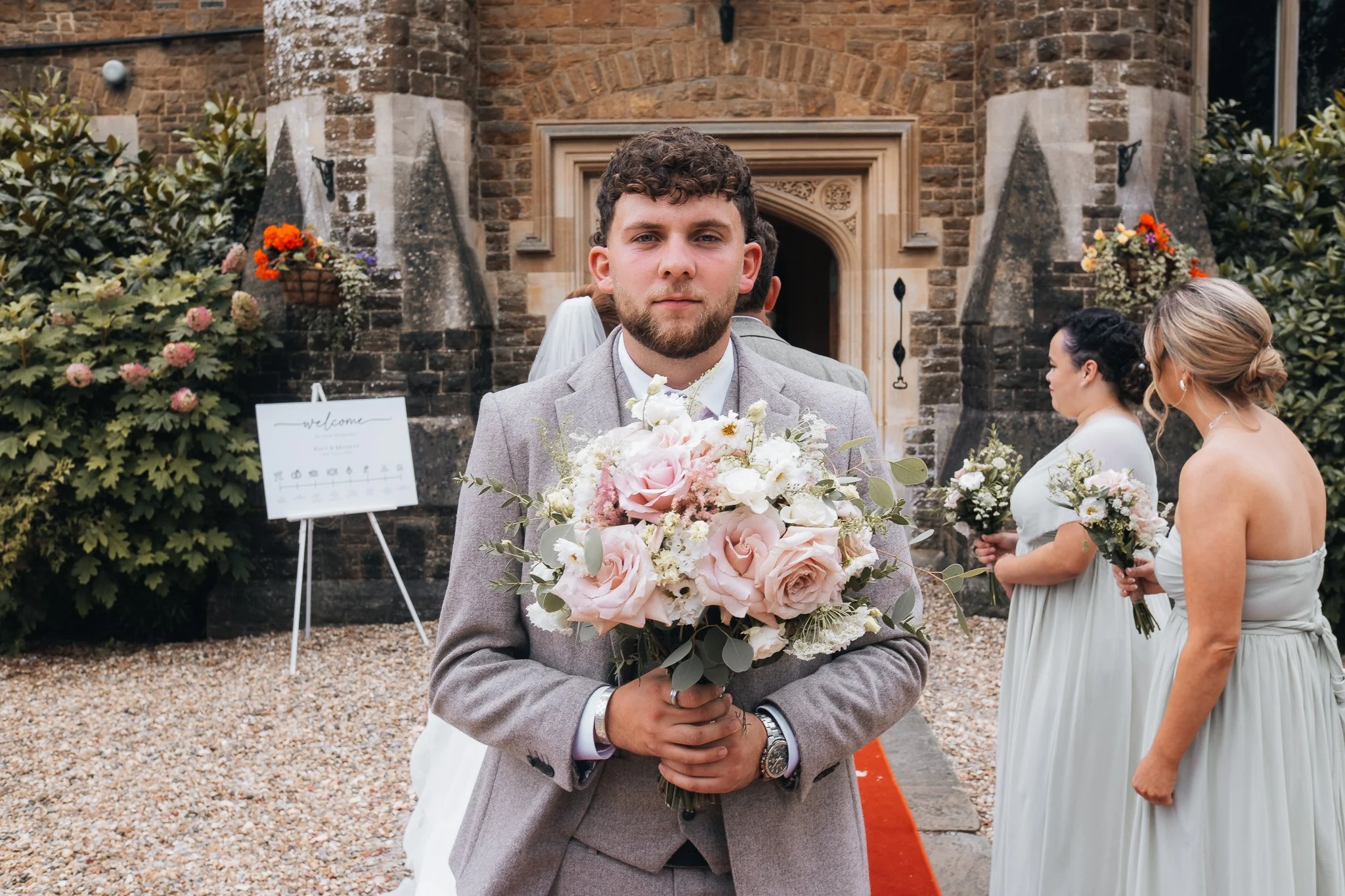 A groom in a light gray suit holding a bouquet of pink and white roses at a wedding ceremony outside a stone church, with bridesmaids in pale dresses standing behind him.