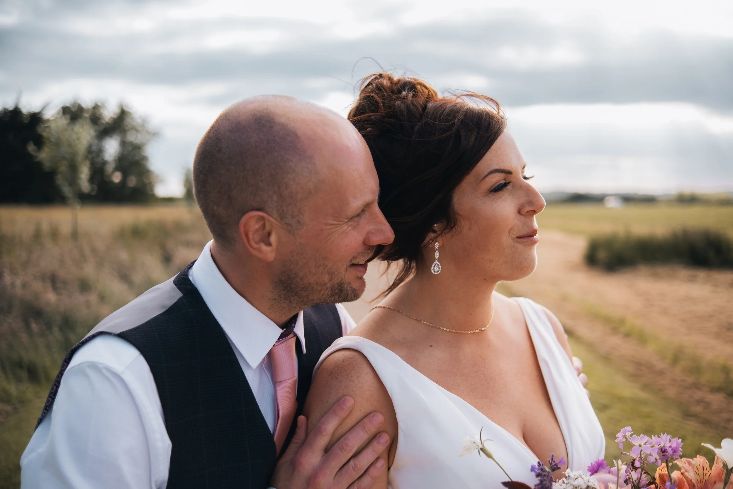 A man and woman in wedding attire outdoors on a cloudy day. The man is speaking quietly into the woman's ear, and both are smiling. The woman is holding a bouquet of flowers.