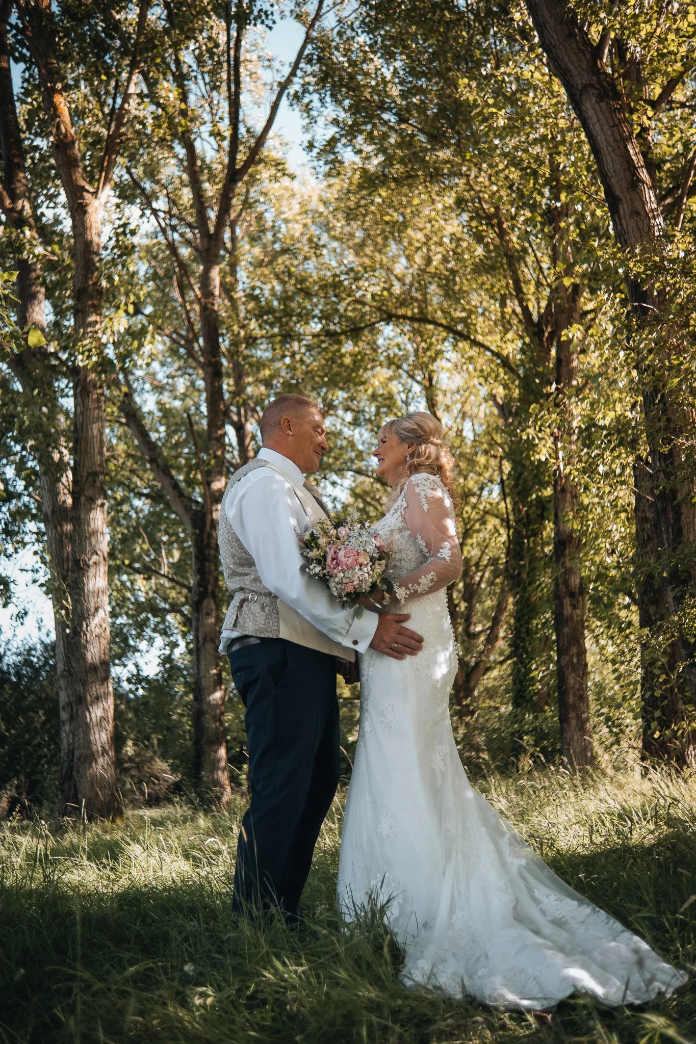 A bride and groom stand close in a forested outdoor setting, sharing an intimate moment, with trees and sunlight overhead.