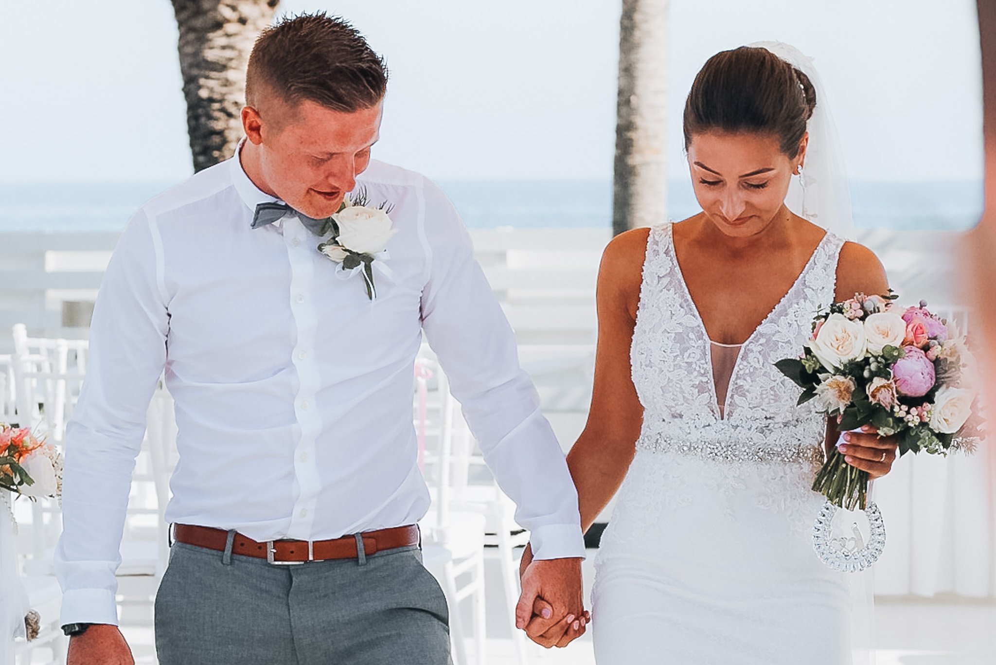 A bride and groom holding hands during their outdoor wedding ceremony on the beach, with palm trees and the ocean in the background.