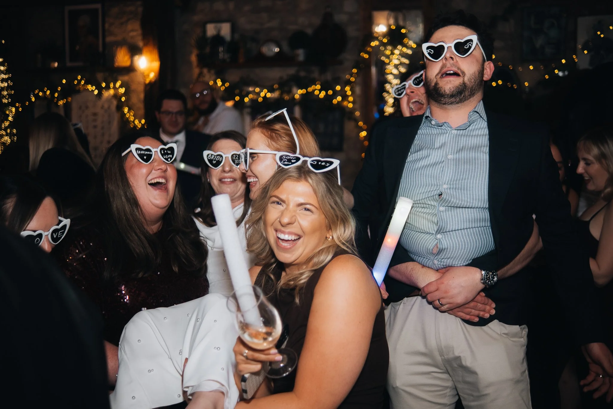 Group of people at a party wearing heart-shaped sunglasses with words like "LOVE" and "BRIDE" on them, some holding glow sticks, in a decorated indoor venue with string lights.
