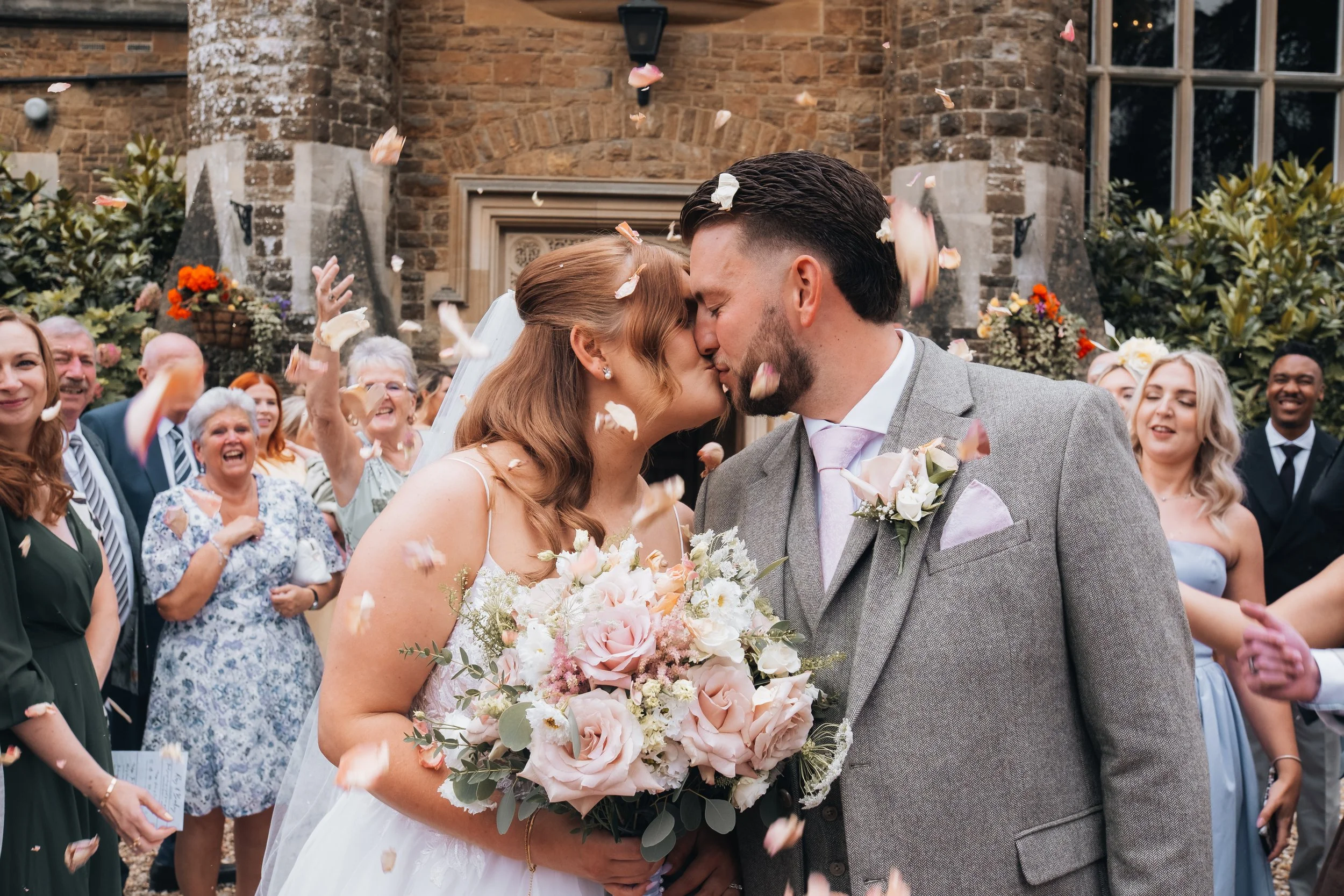 A newlywed couple kissing amidst flower petal-throwing guests outside a stone church.
