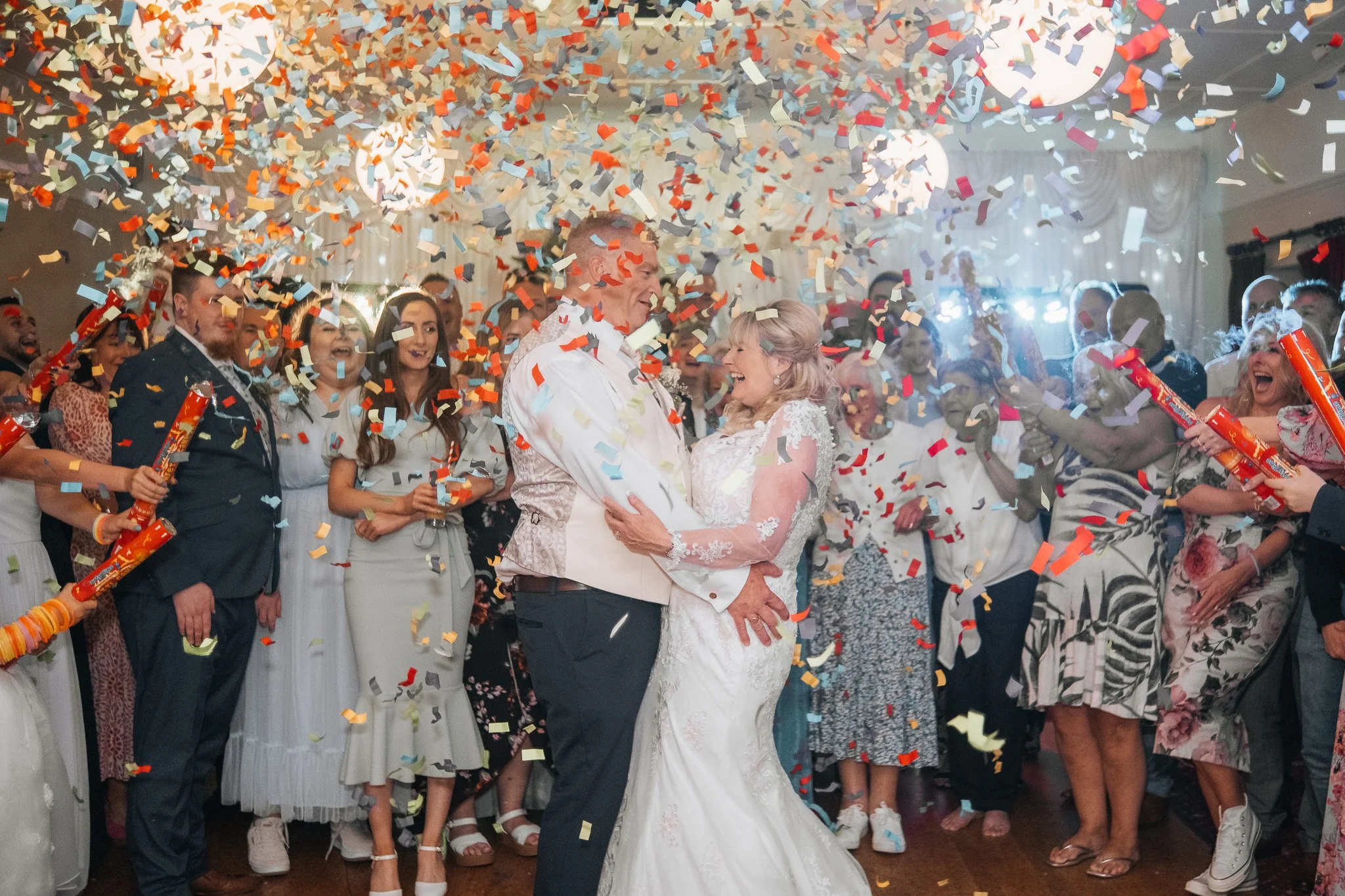 Happy bride and groom dancing amidst shower of colorful confetti at their wedding reception, surrounded by smiling guests