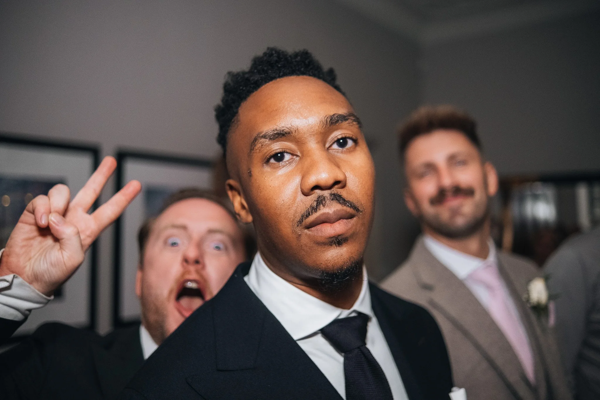 Three men in suits at a social gathering, with one man in the foreground making a serious expression, while the other two men behind him are smiling, one making a peace sign.