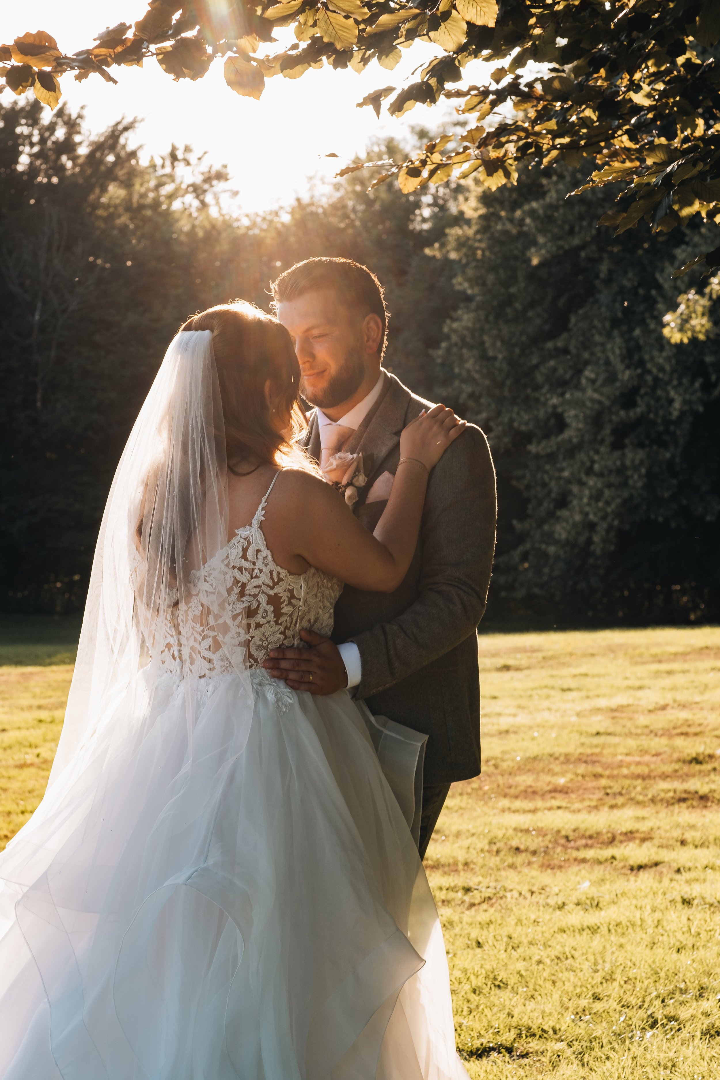 A bride and groom share a romantic dance outdoors at sunset, with trees in the background and sunlight shining through leaves.