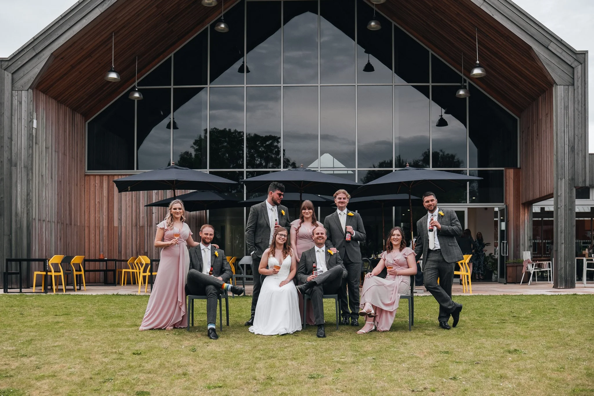 Group of wedding guests, some seated and some standing, posing outdoors in front of a modern barn-style building with large glass windows and black umbrellas, on a cloudy day.