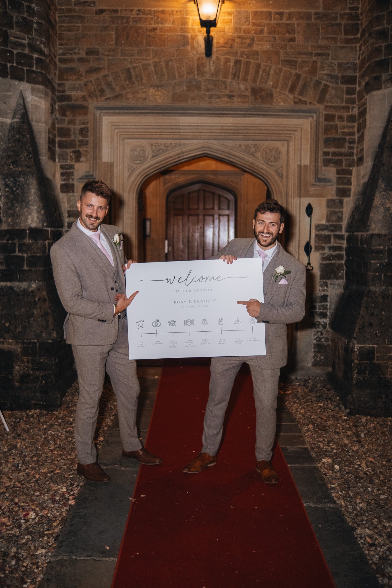 Two men, dressed in grey suits with pink ties and white shirts, standing on a red carpet in front of an arched doorway with a lantern above, holding a large welcome sign for their wedding.