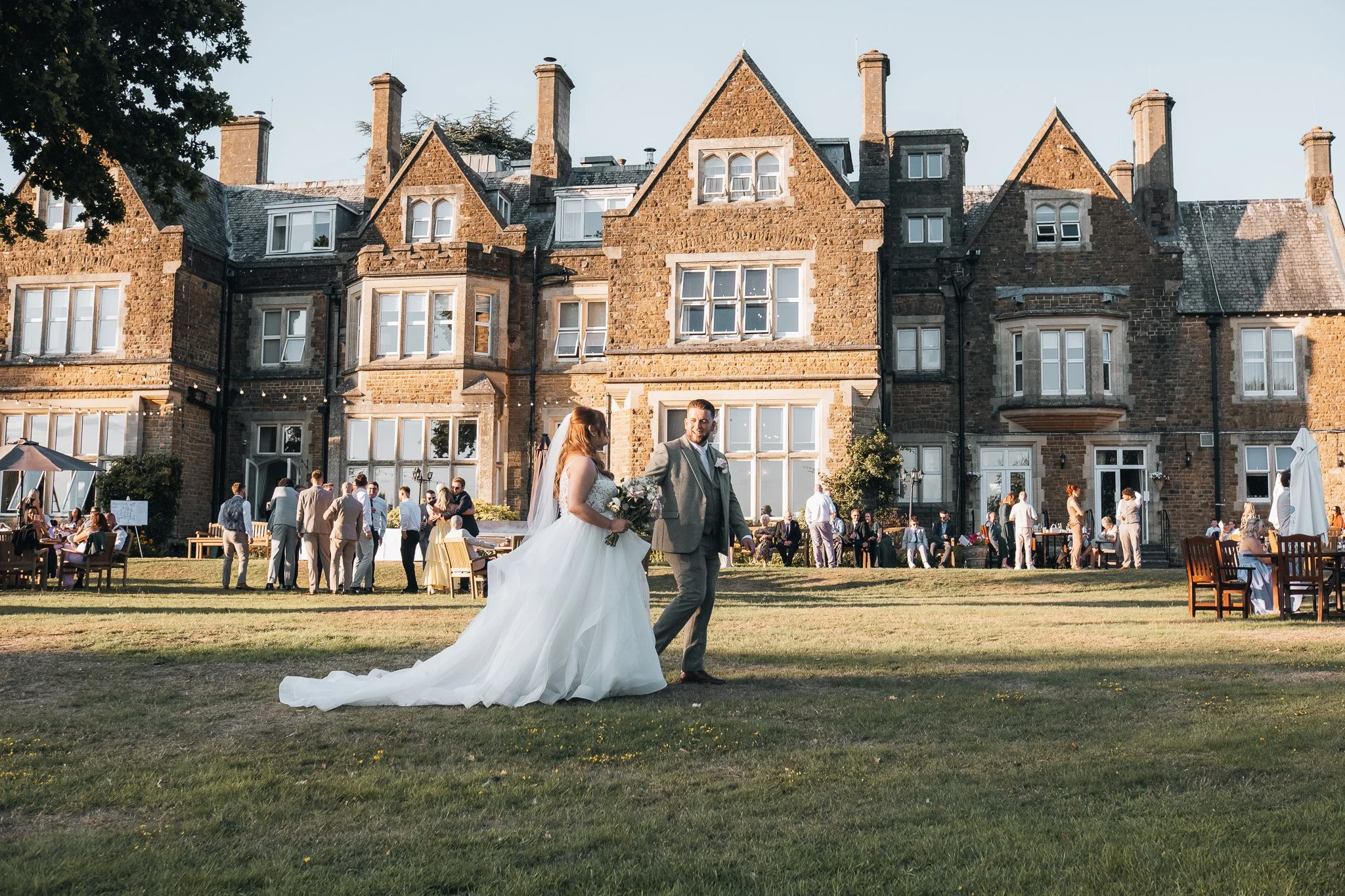 A bride and groom are walking hand-in-hand across a grassy lawn in front of a large historic brick building during a wedding celebration. The bride is in a white wedding dress holding a bouquet, and the groom is in a gray suit. Guests are gathered an