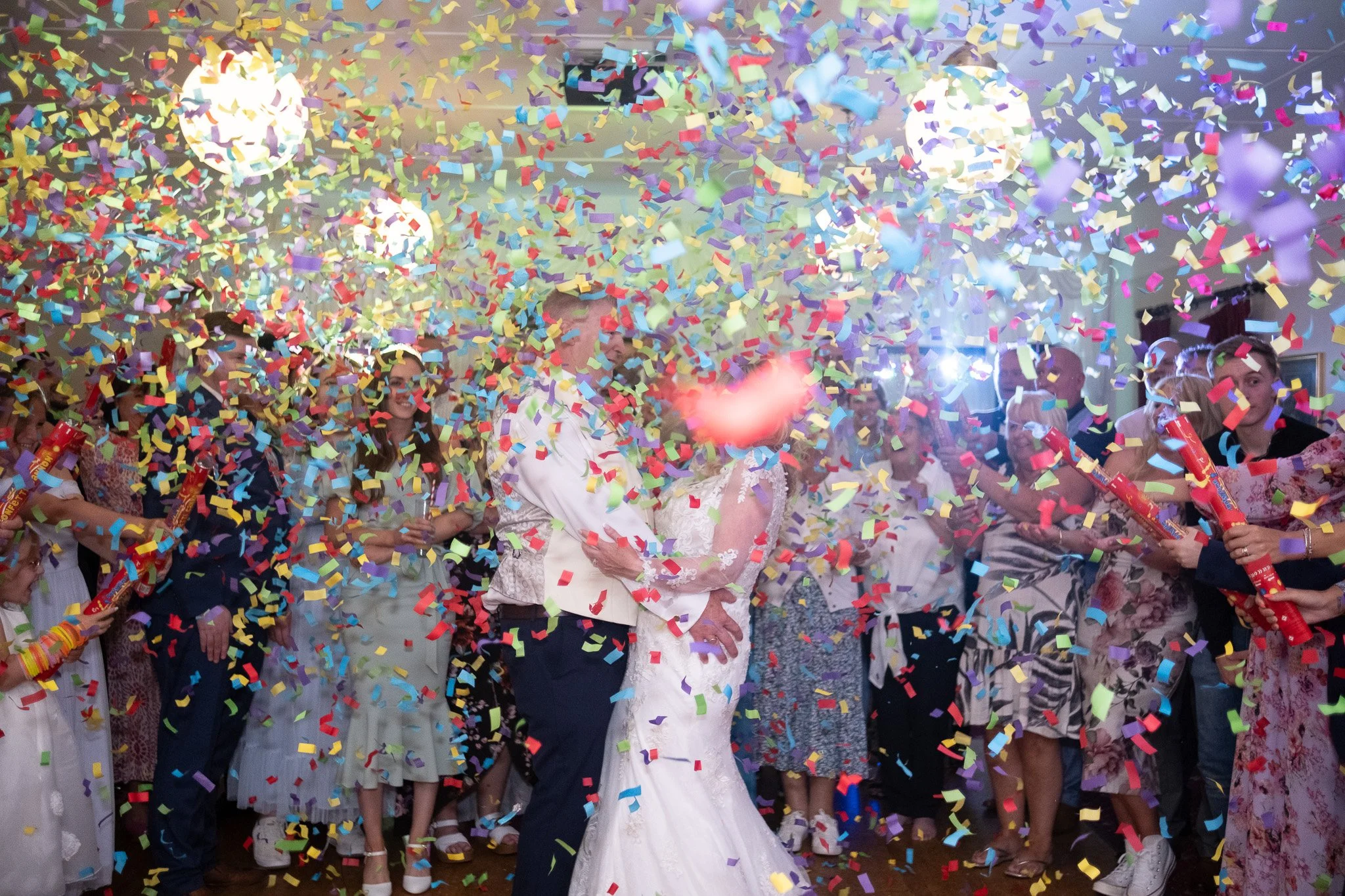 Couple kissing under falling colorful confetti at celebration with guests surrounding them in festive attire.