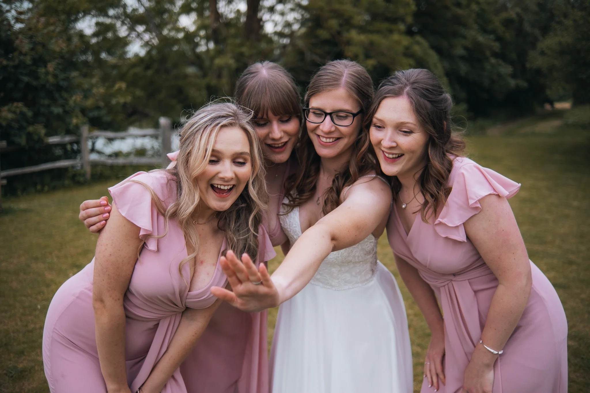 Four women, one in a wedding dress and three bridesmaids in pink dresses, smiling and looking at something they are holding or pointing to outside in a park.