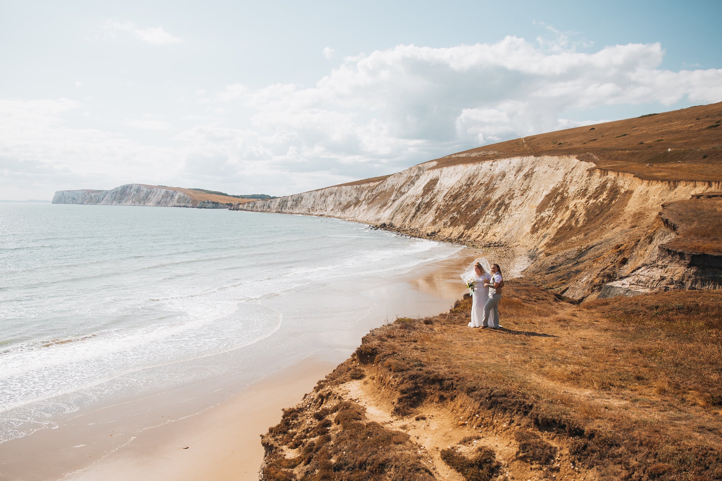 A bride and groom standing on a cliffside beach with white chalk cliffs in the background, under a partly cloudy sky.