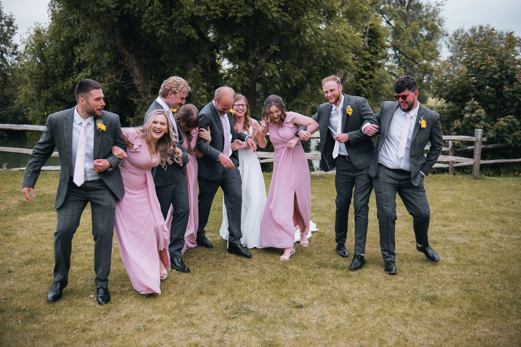 Group of wedding guests, including men in suits and women in pink dresses, walking and laughing together on a grassy outdoor area with trees and a wooden fence in the background.