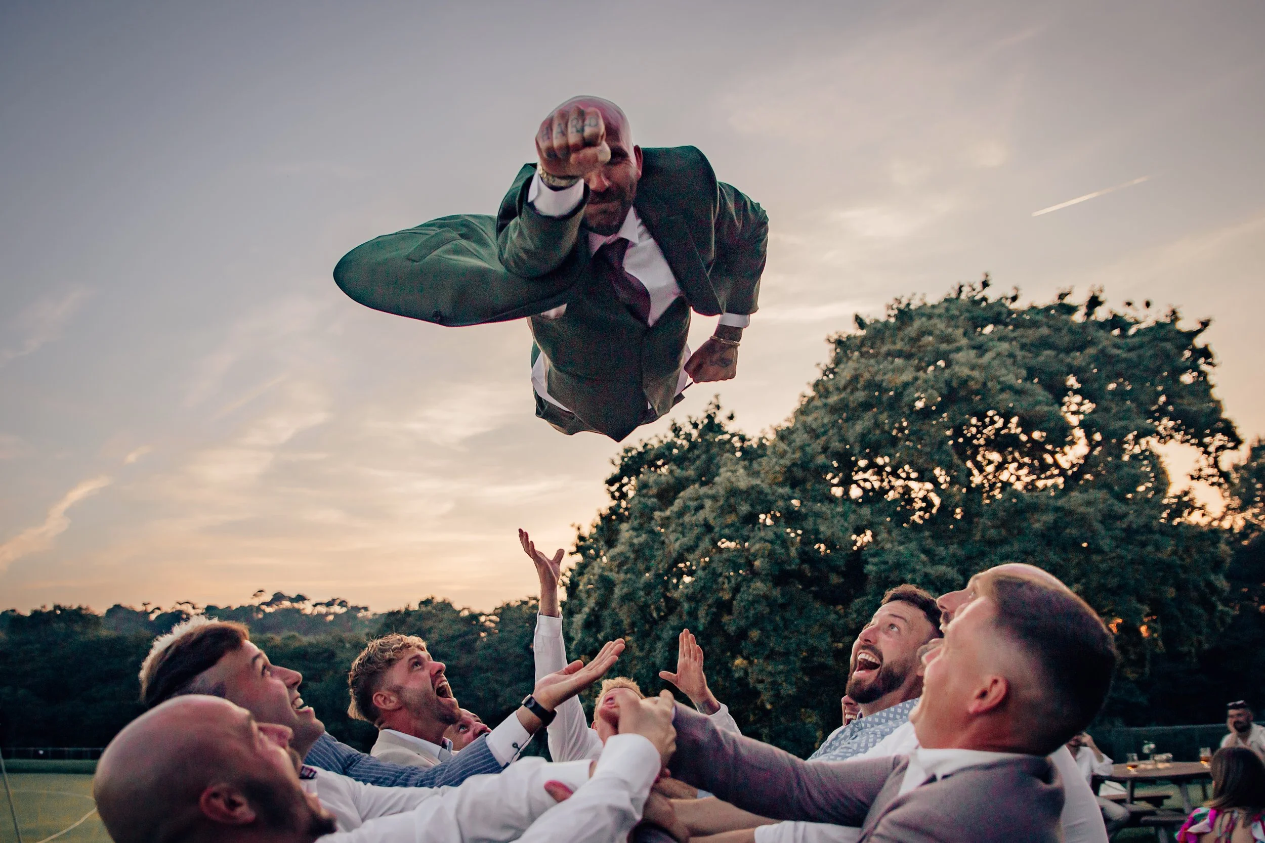 A man in a suit is flying through the air, being tossed by a group of people outdoors during sunset, with trees in the background and people looking up and cheering.