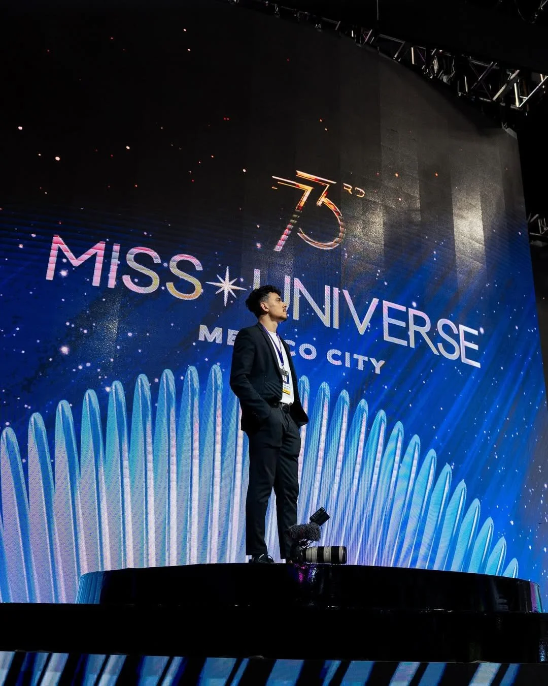 Hombre de negocios con traje en el escenario durante la celebración del 75 aniversario de Miss Universe en Ciudad de México, con fondo de pantalla con temática espacial y letras brillantes.